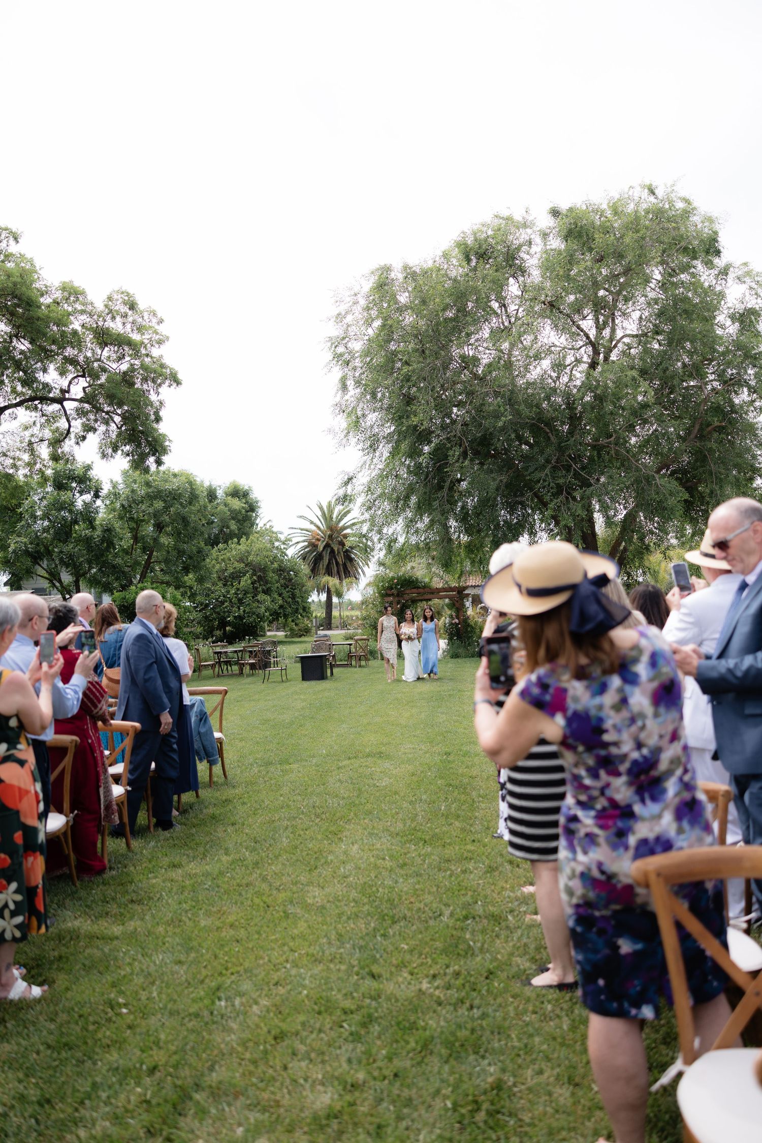Guests stand and watch as the bride walks down the aisle at an outdoor garden wedding ceremony.