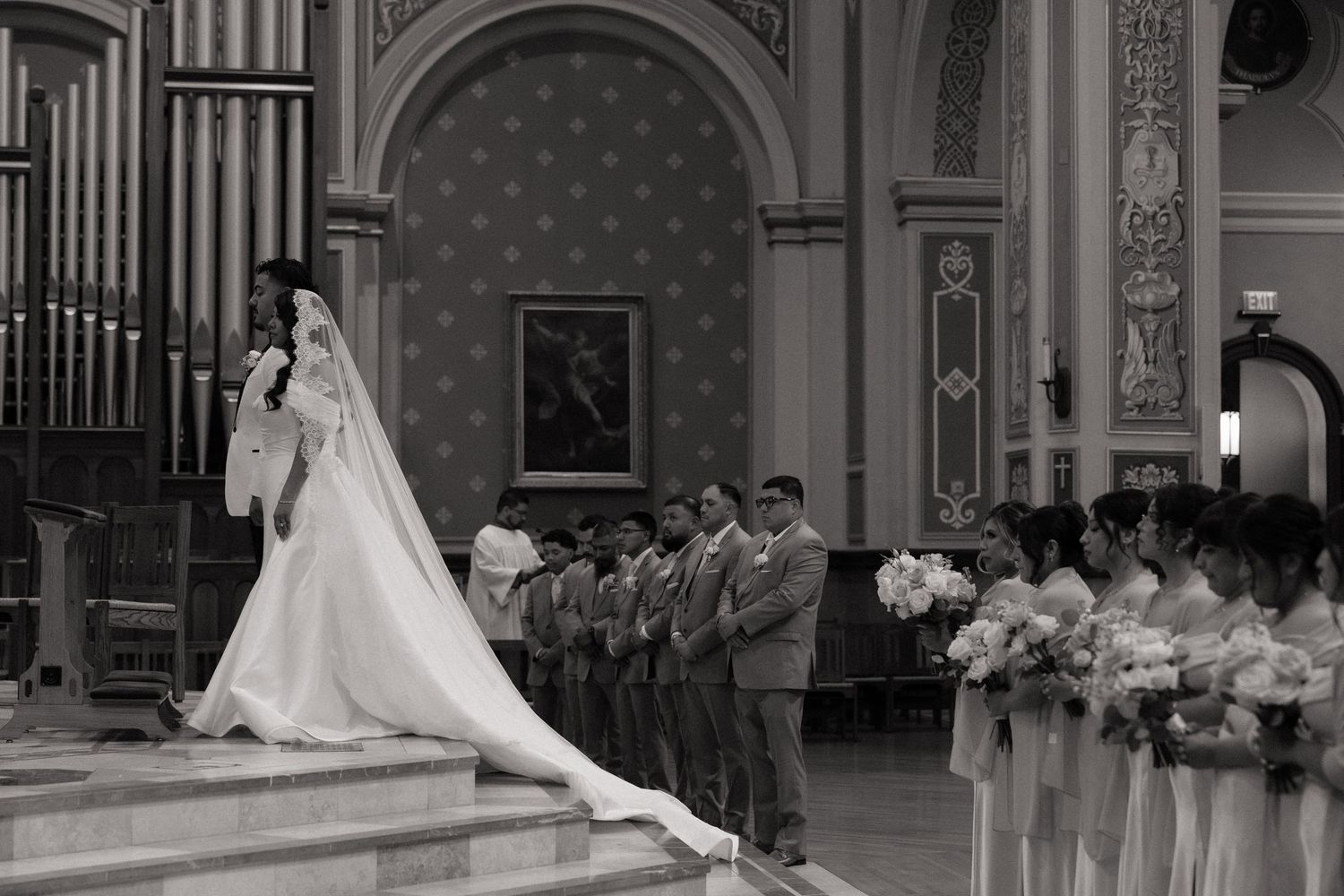 Bride in elegant gown stands at church altar with bridesmaids and groomsmen during wedding ceremony.