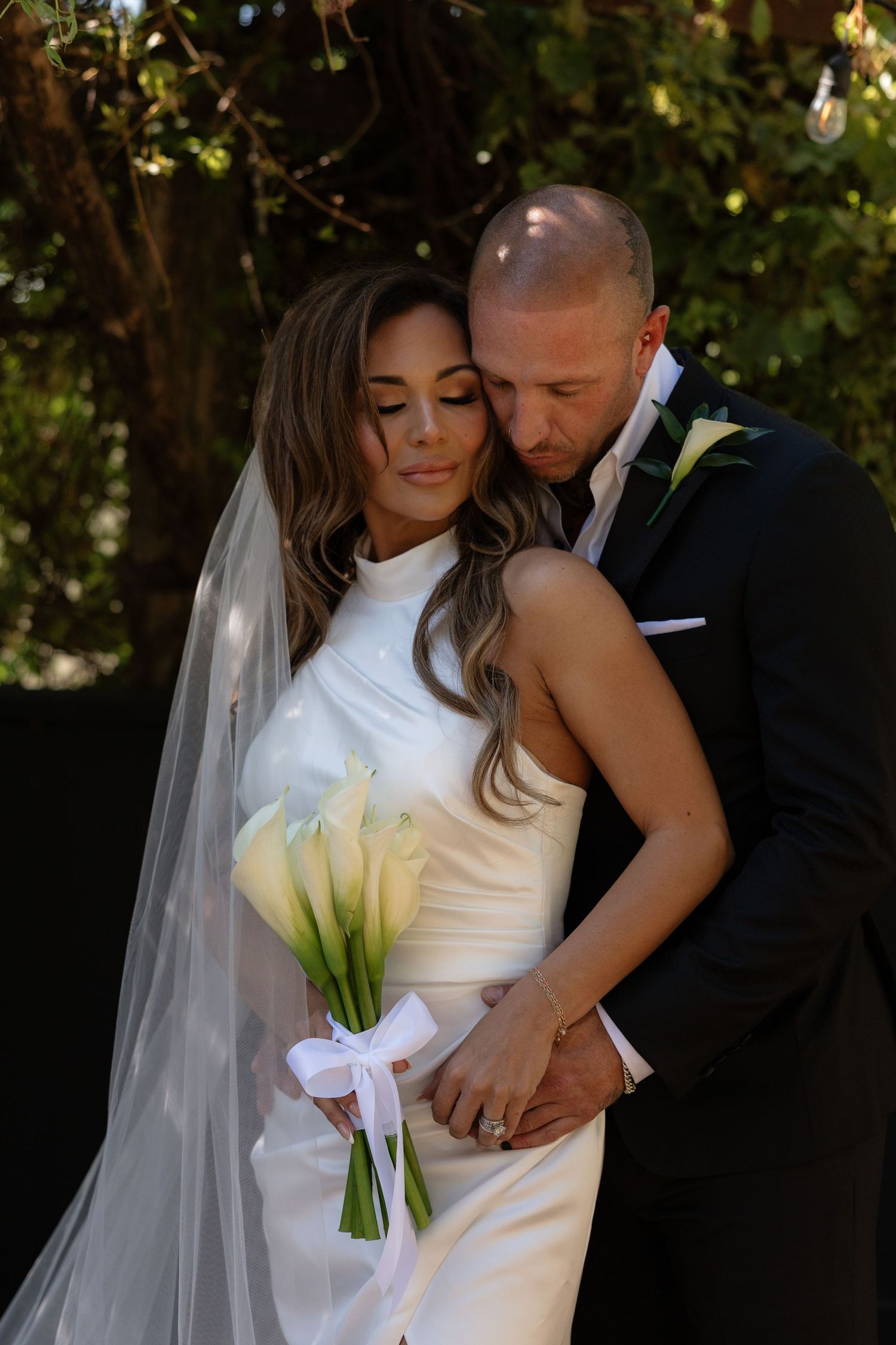 Bride in white gown holding calla lily bouquet embraced by groom in black suit at outdoor wedding ceremony.