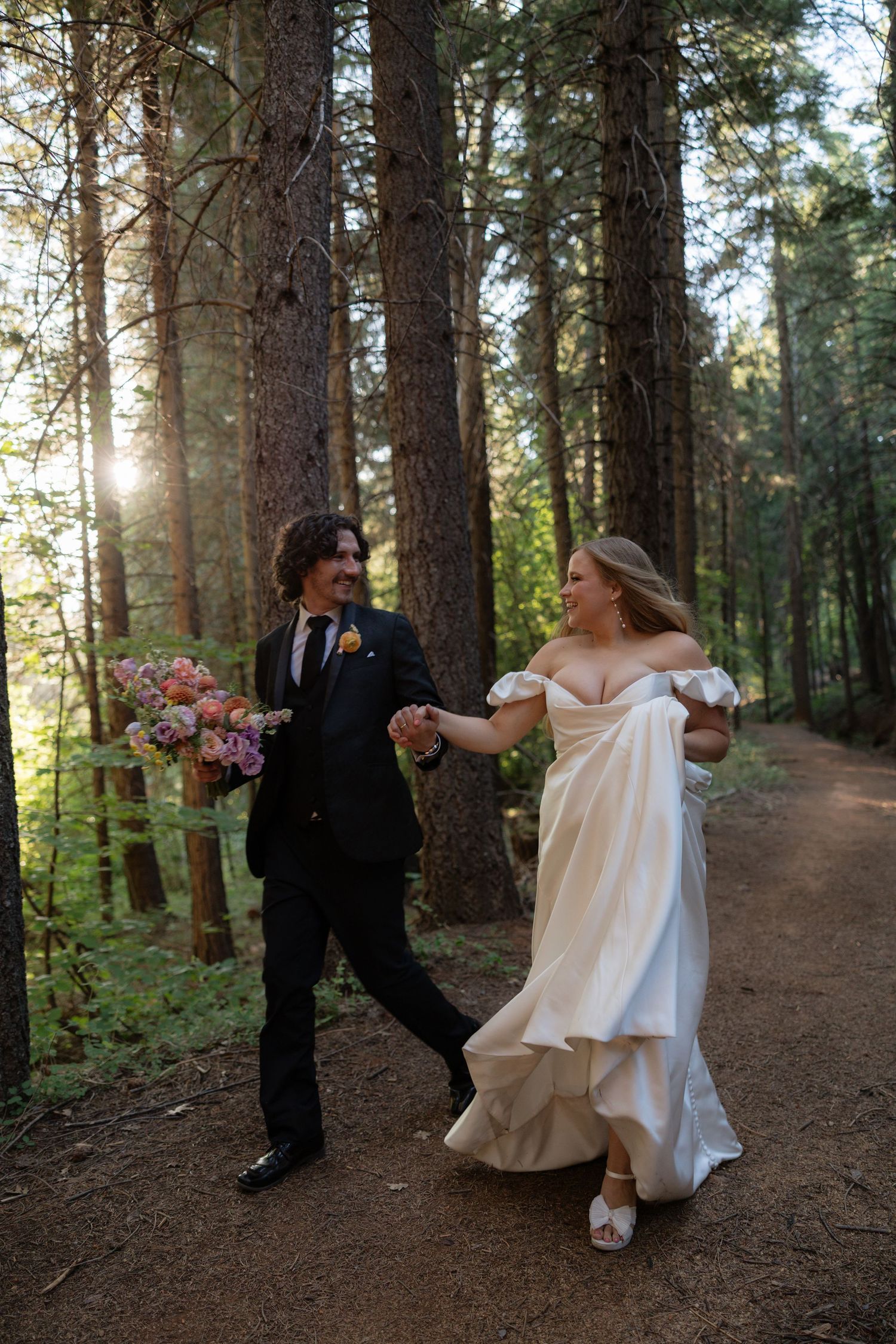 Bride and groom walking hand-in-hand through a sunlit forest, bride in white gown holding pink bouquet.