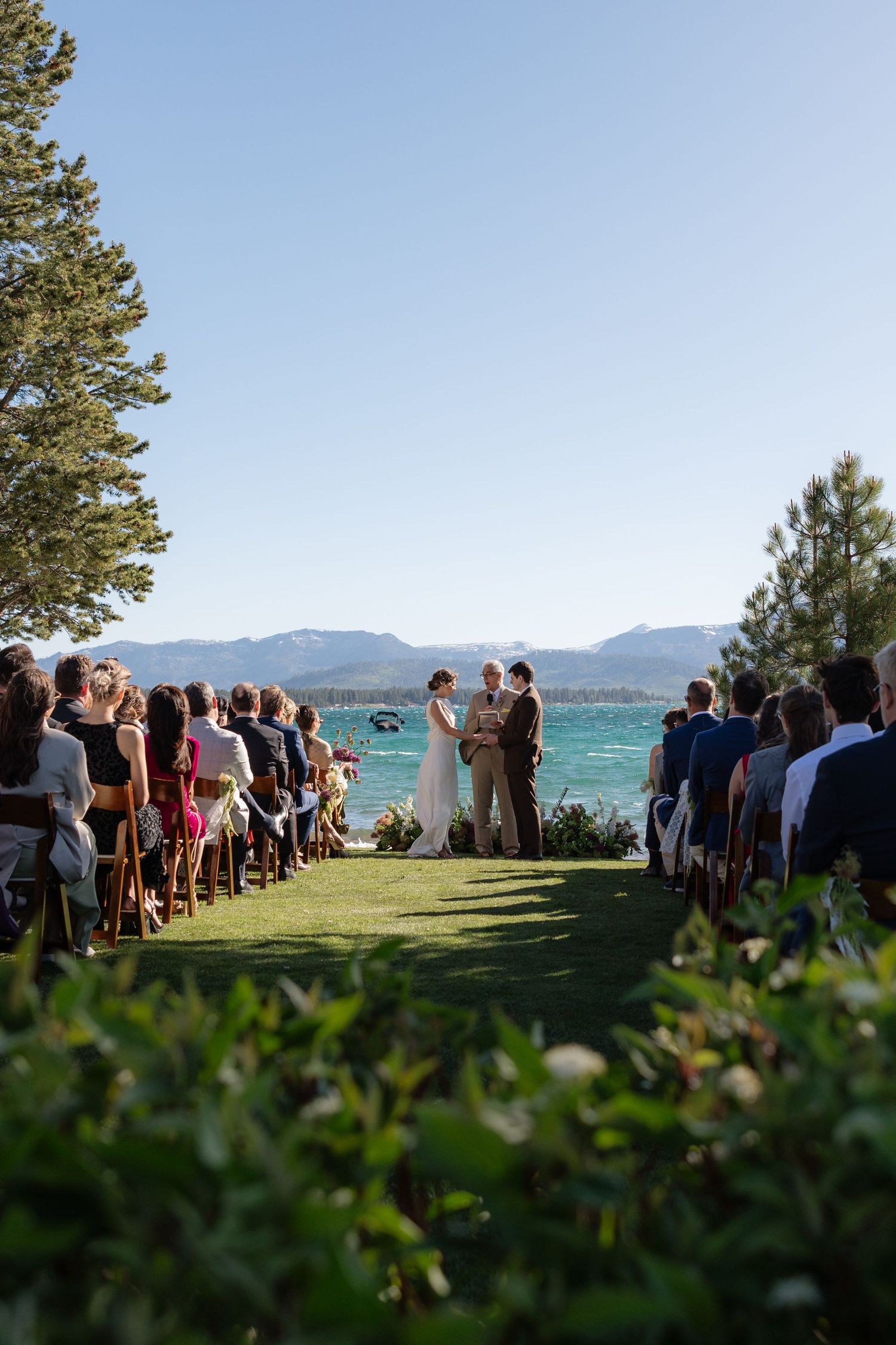 Outdoor wedding ceremony on green lawn with lake and mountain backdrop under clear blue sky.