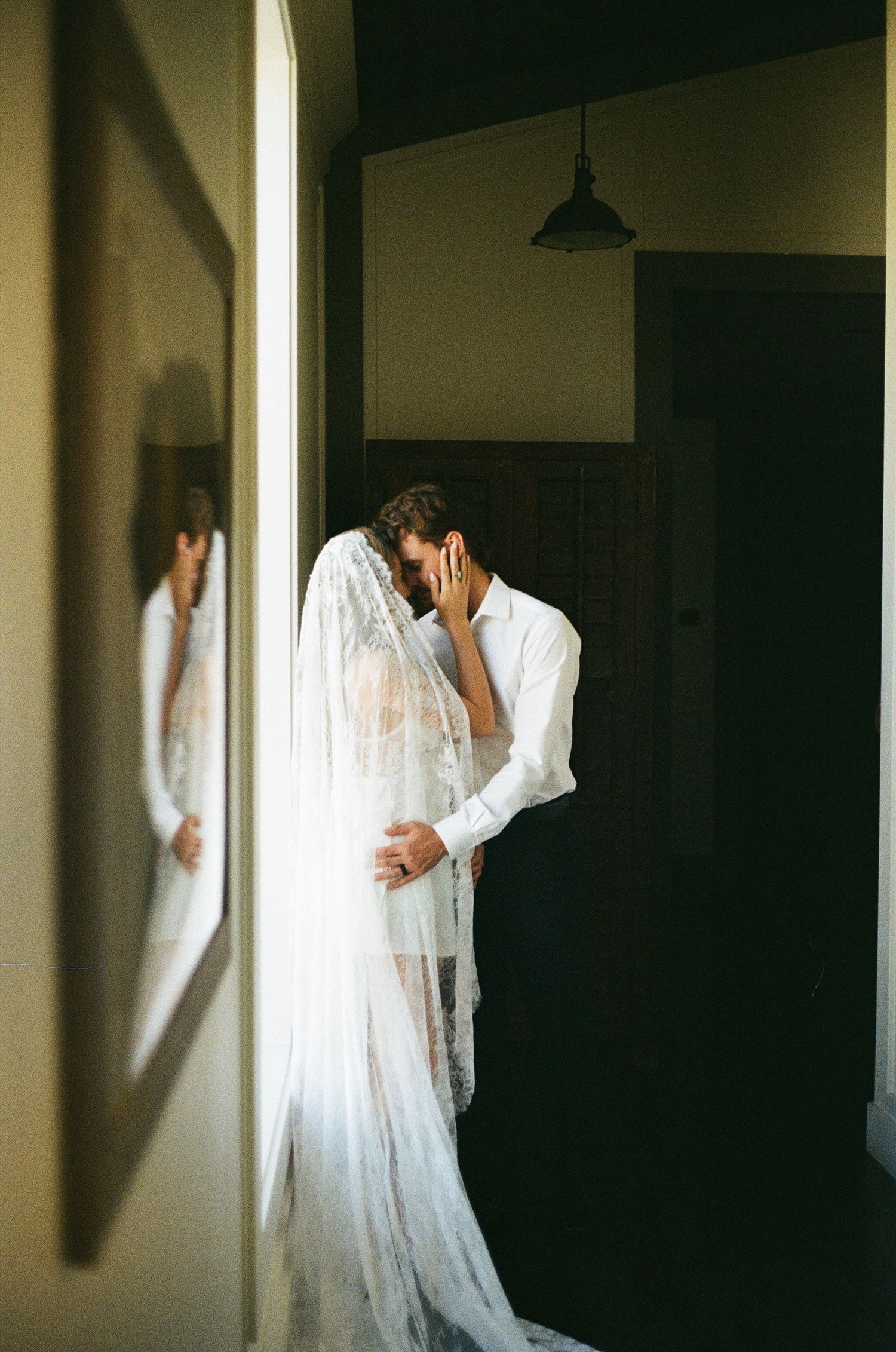 A bride in a lace veil and gown embraces her groom in a white shirt, sharing an intimate moment indoors.
