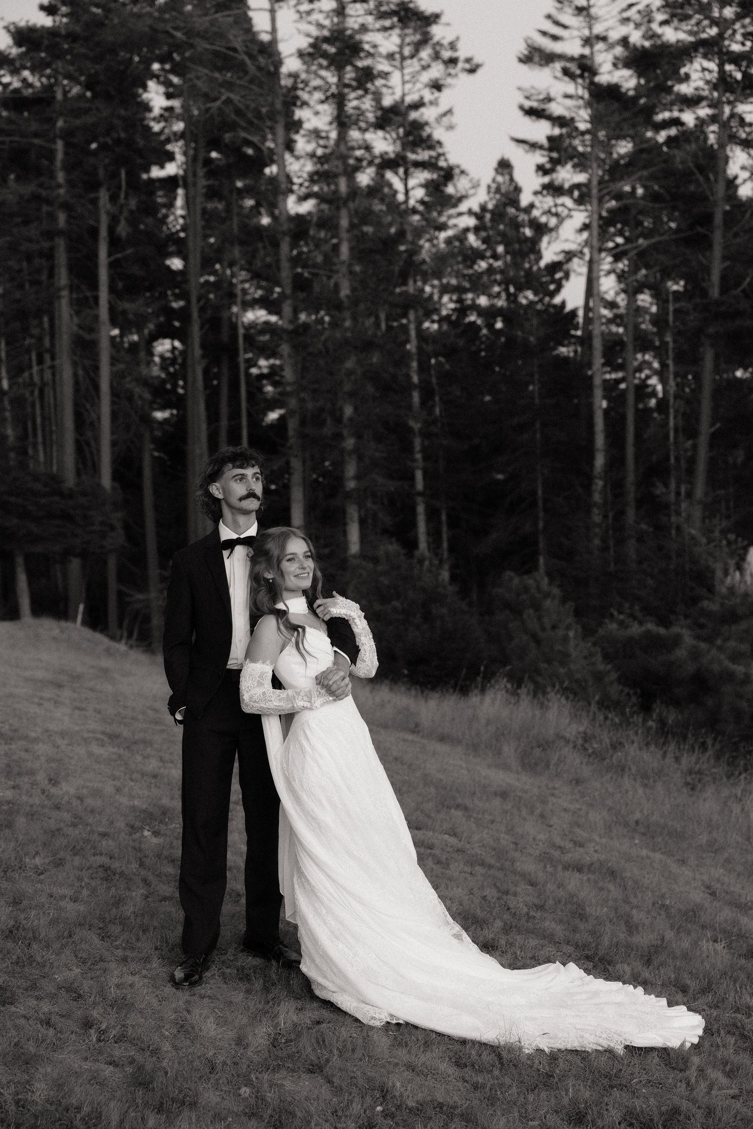 Black and white photo of a bride and groom embracing in a forest clearing, groom in tuxedo, bride in flowing white gown.
