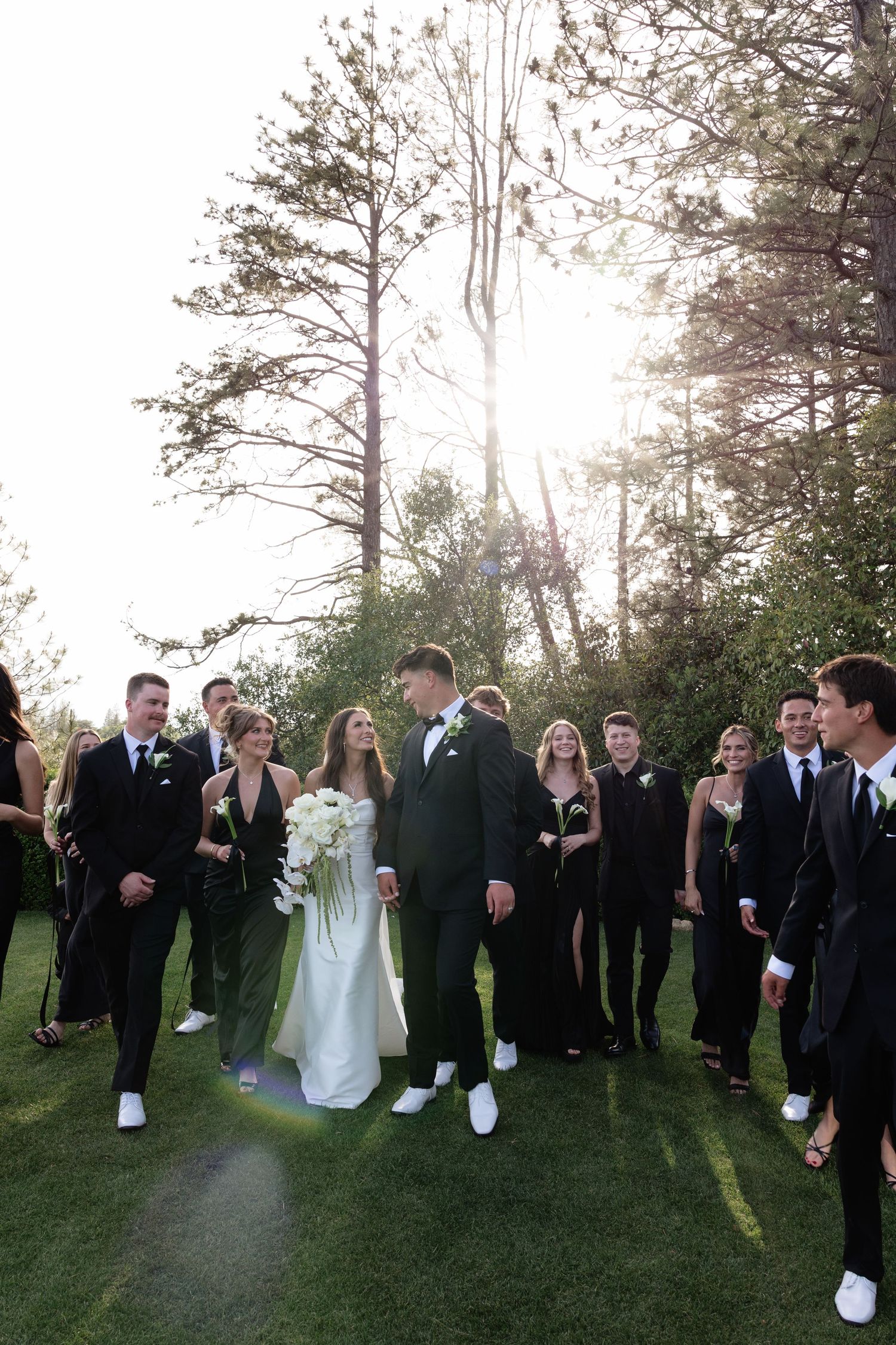 Wedding party walks outdoors in black attire with bride in white gown holding bouquet, sunlight filtering through trees.