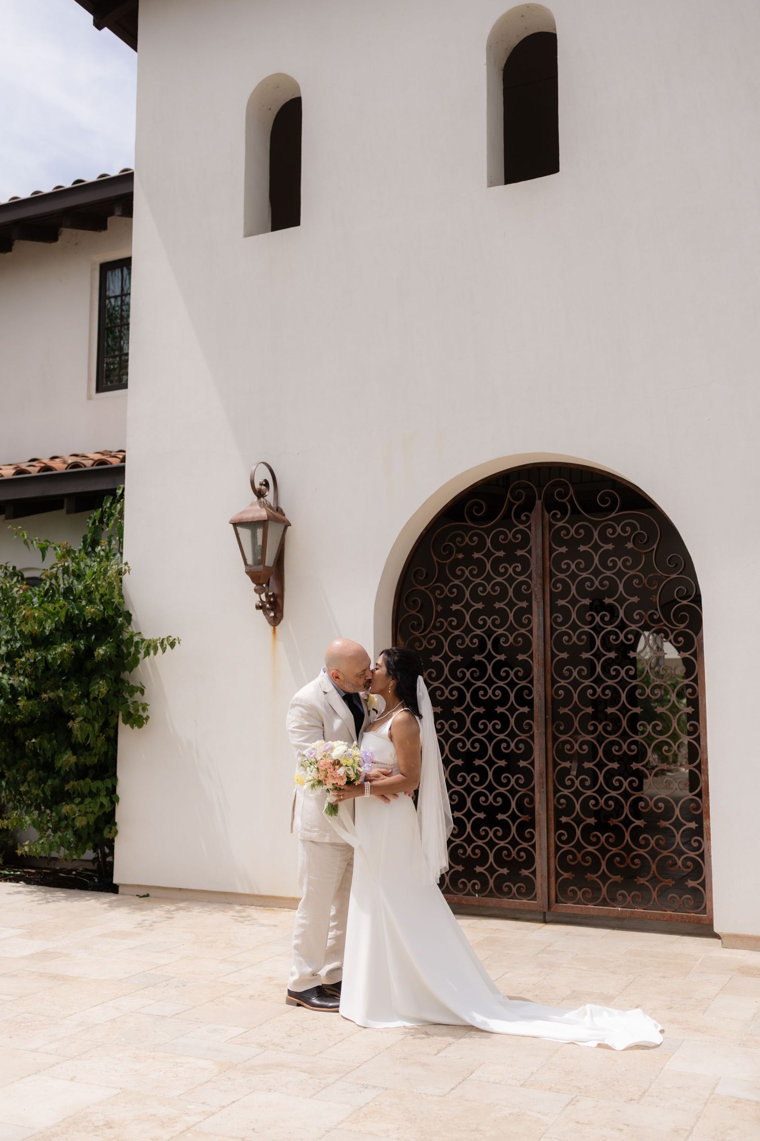 Bride and groom share a kiss outside a Spanish-style white stucco building with ornate iron doors.