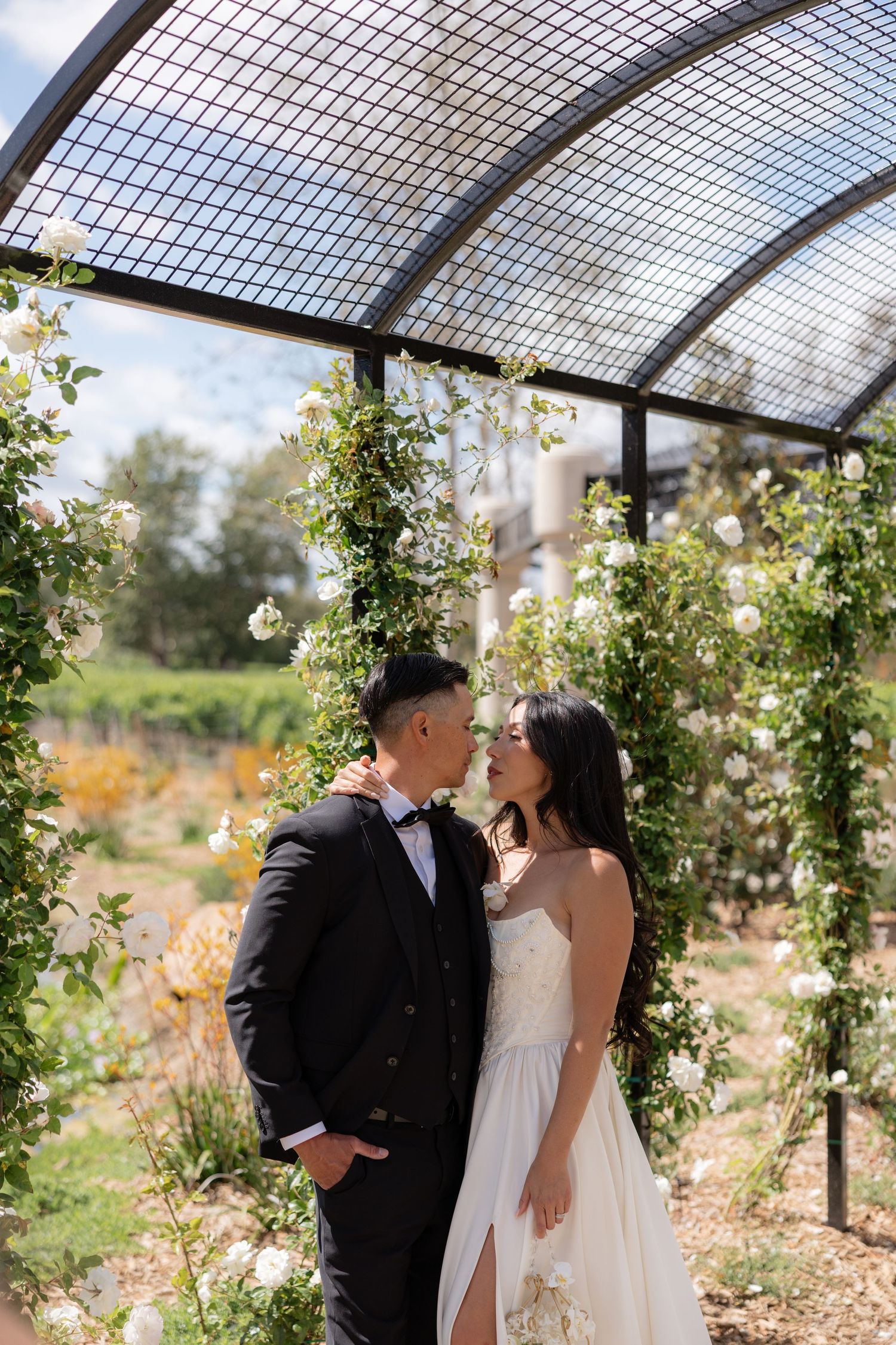 Bride and groom sharing a romantic moment under a white rose-covered garden arch at their wedding.