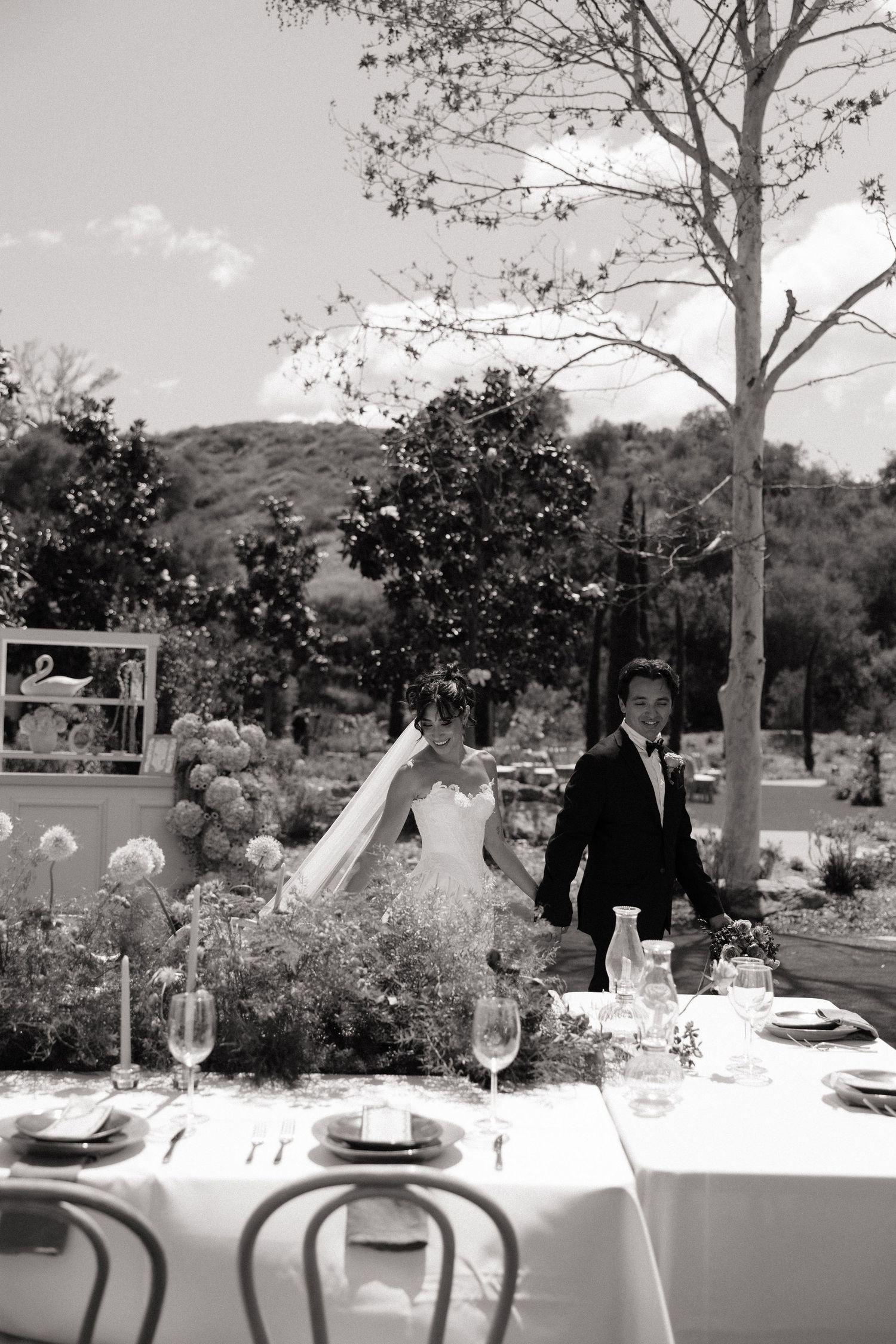 Bride and groom strolling past an elegantly set outdoor reception table in black and white photo.