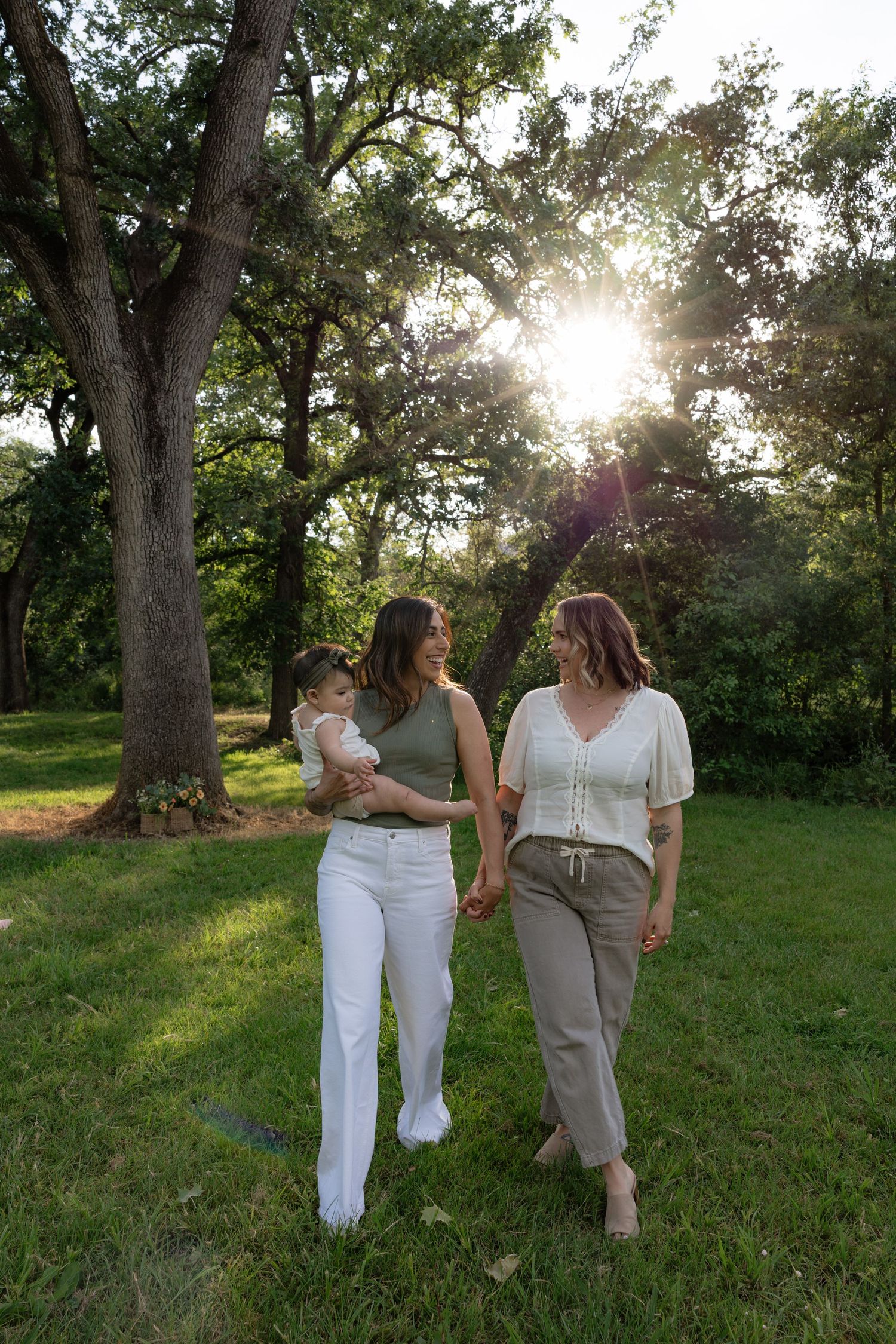 Two women holding hands and carrying a baby while walking in a sunny green park.