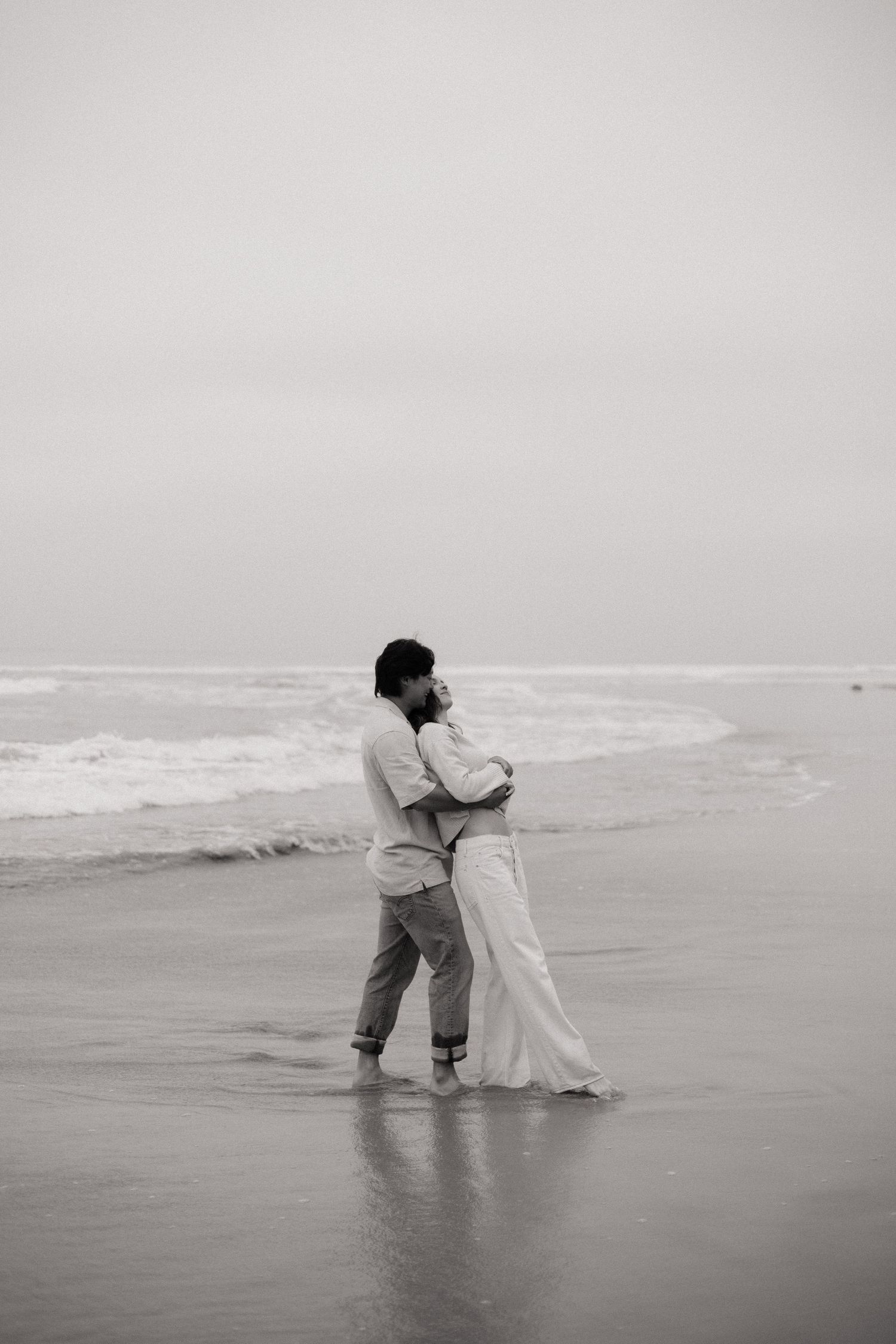 Black and white photo of a couple embracing on a misty beach, waves rolling in behind them.
