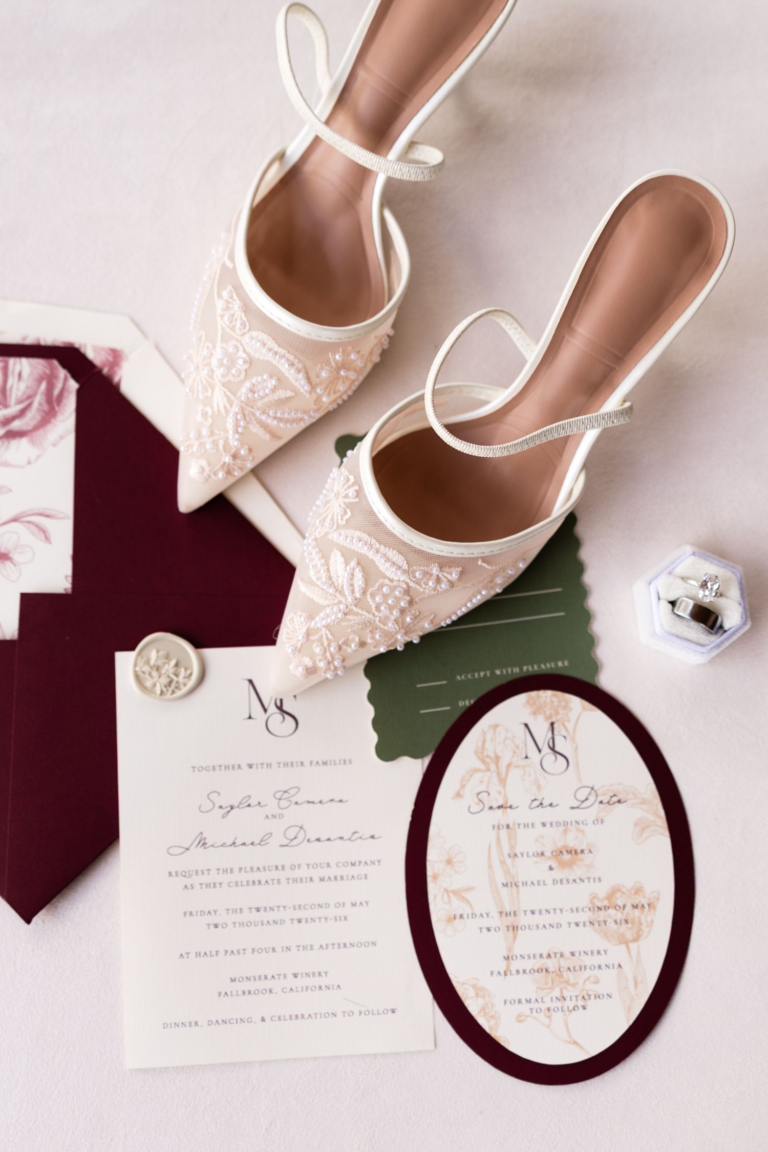 Elegant bridal flat lay featuring lace mule heels, burgundy velvet wedding invitations, wax seal, and diamond ring box.