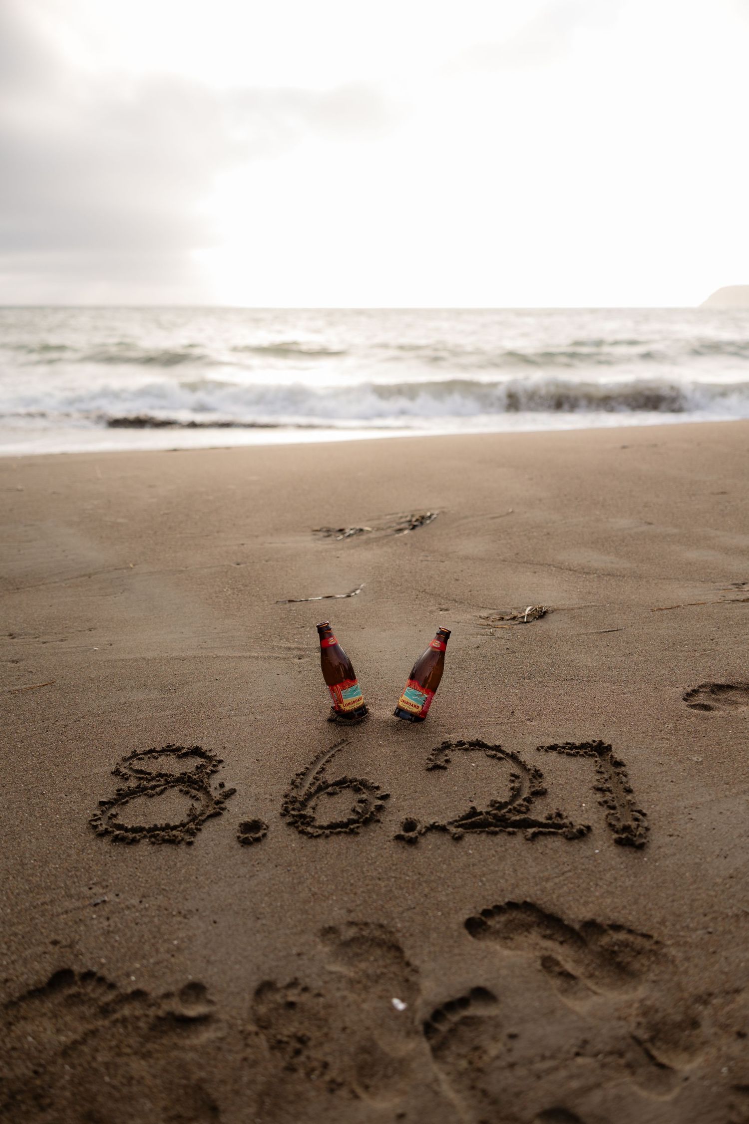 Date 8.6.21 written in beach sand with two beer bottles marking a romantic anniversary celebration at sunset.