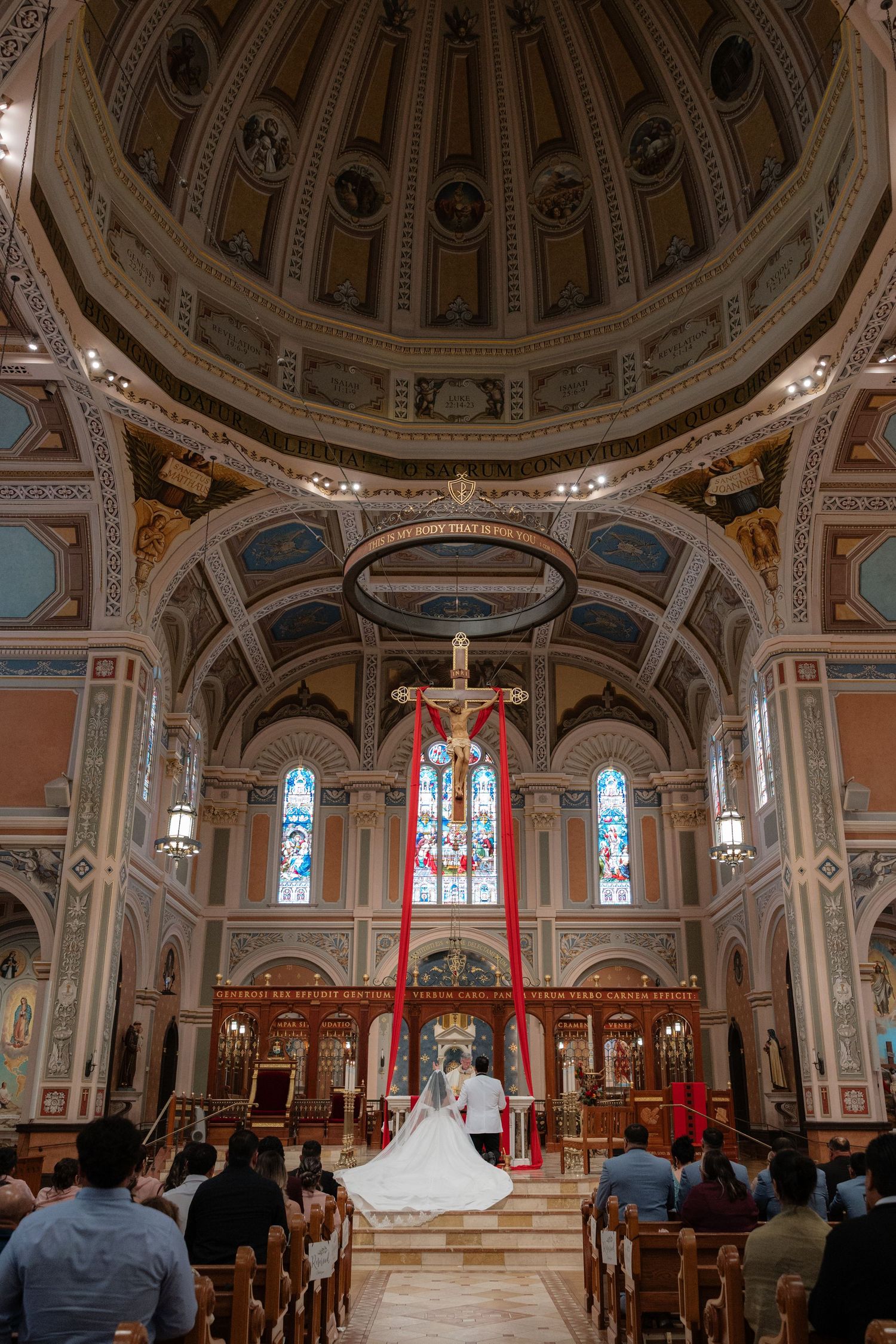 Wedding ceremony inside ornate cathedral with domed ceiling, stained glass windows, and crucifix altar.