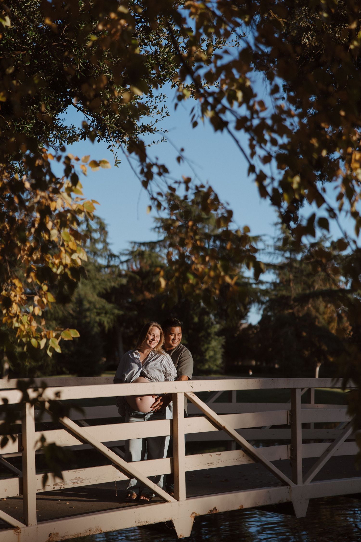 Couple standing on a white bridge surrounded by autumn foliage in a park.