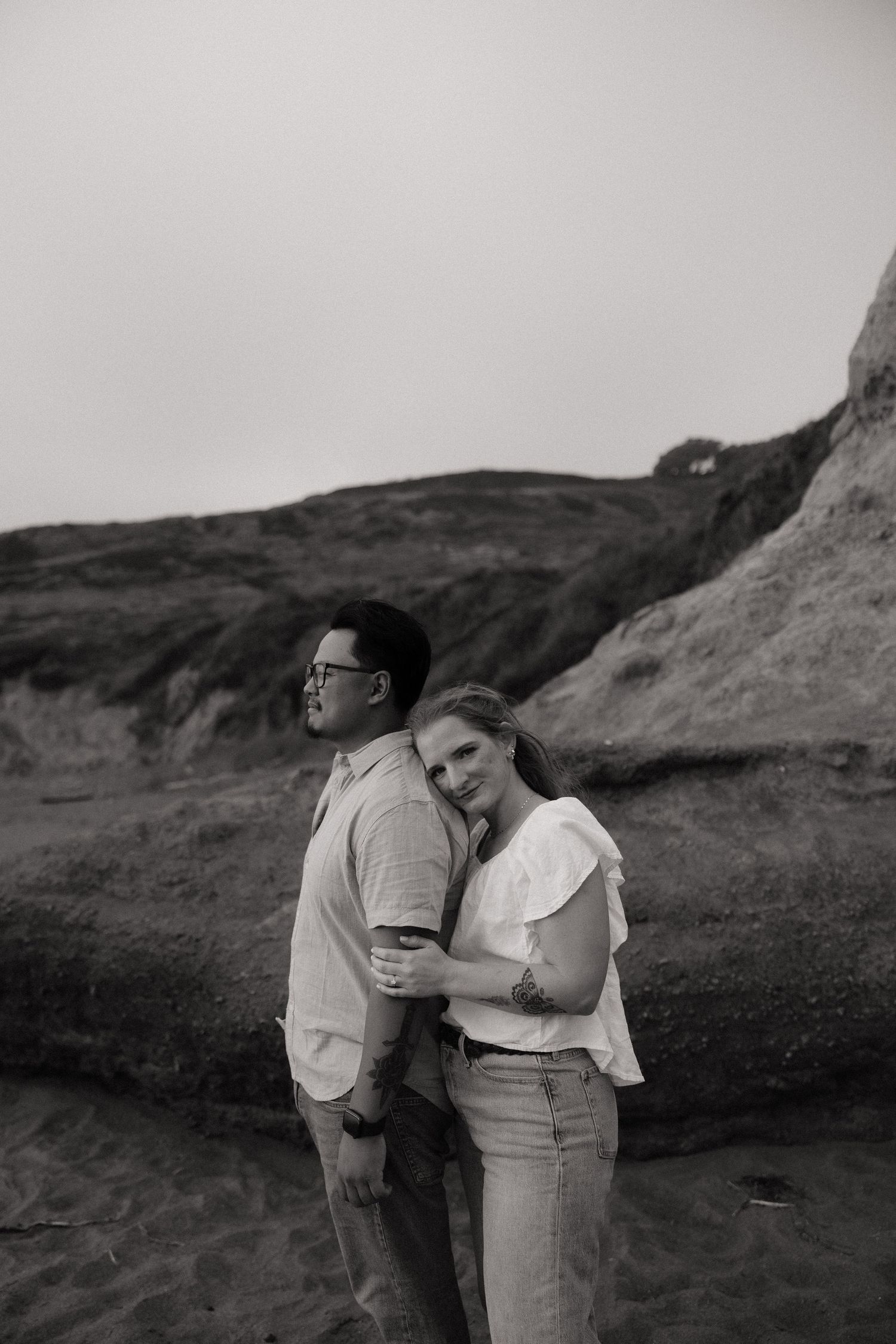 Black and white photo of a couple embracing on a rocky beach, woman resting her head on mans shoulder.