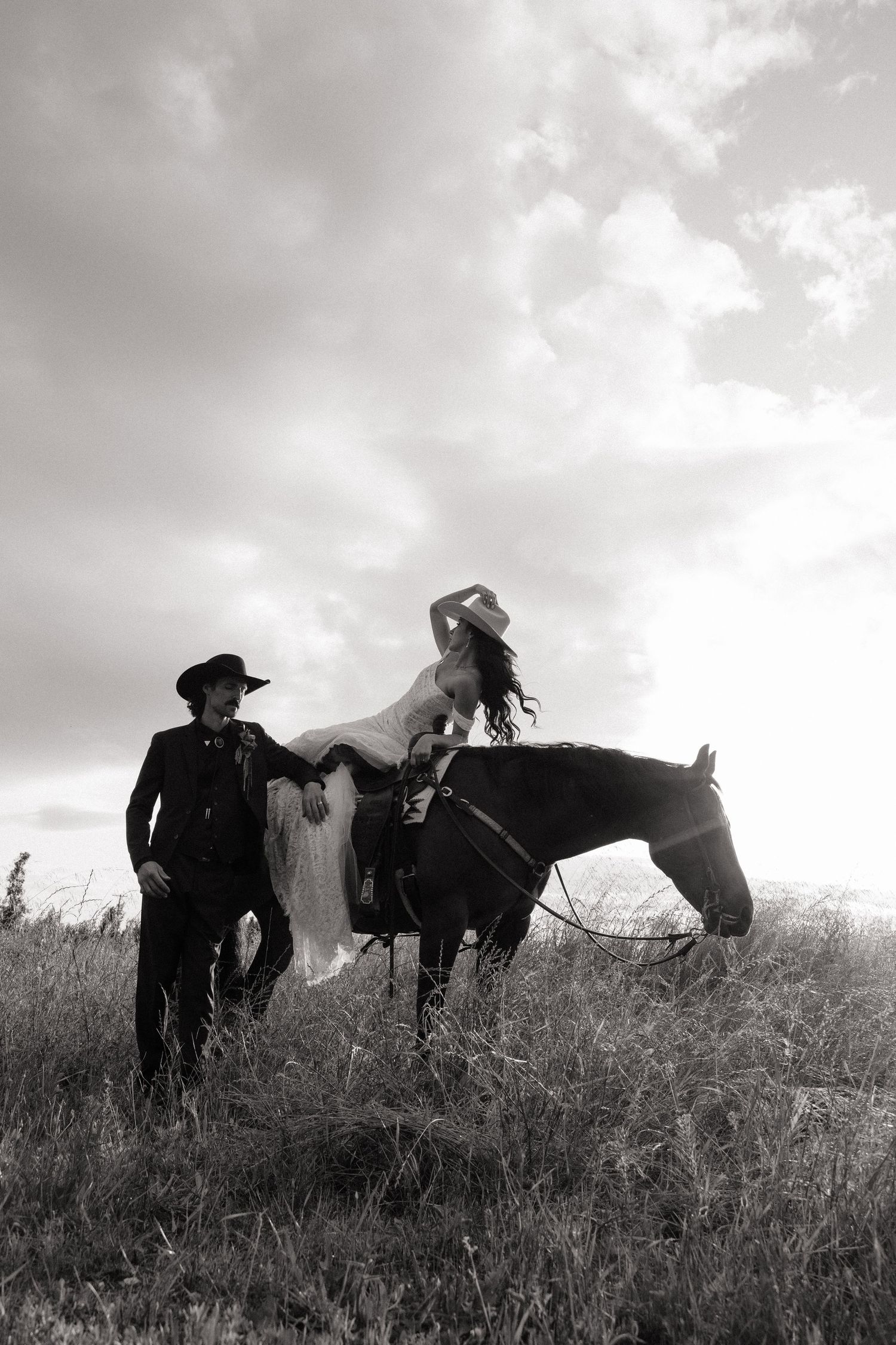 Bride in white gown sitting on horseback with groom in black cowboy hat in an open field.