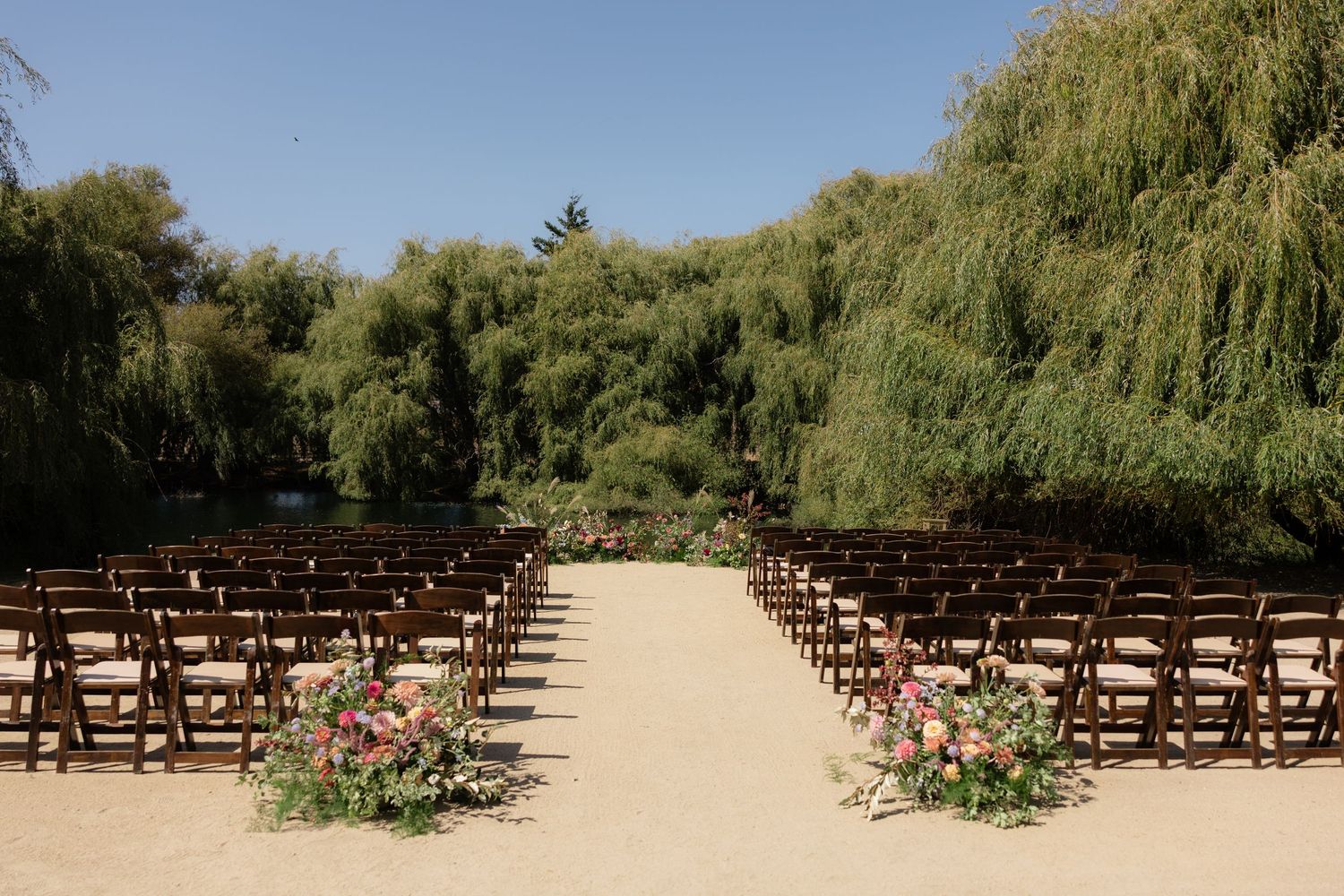 Outdoor wedding ceremony setup with wooden chairs, floral arrangements, and willow trees under a clear blue sky.