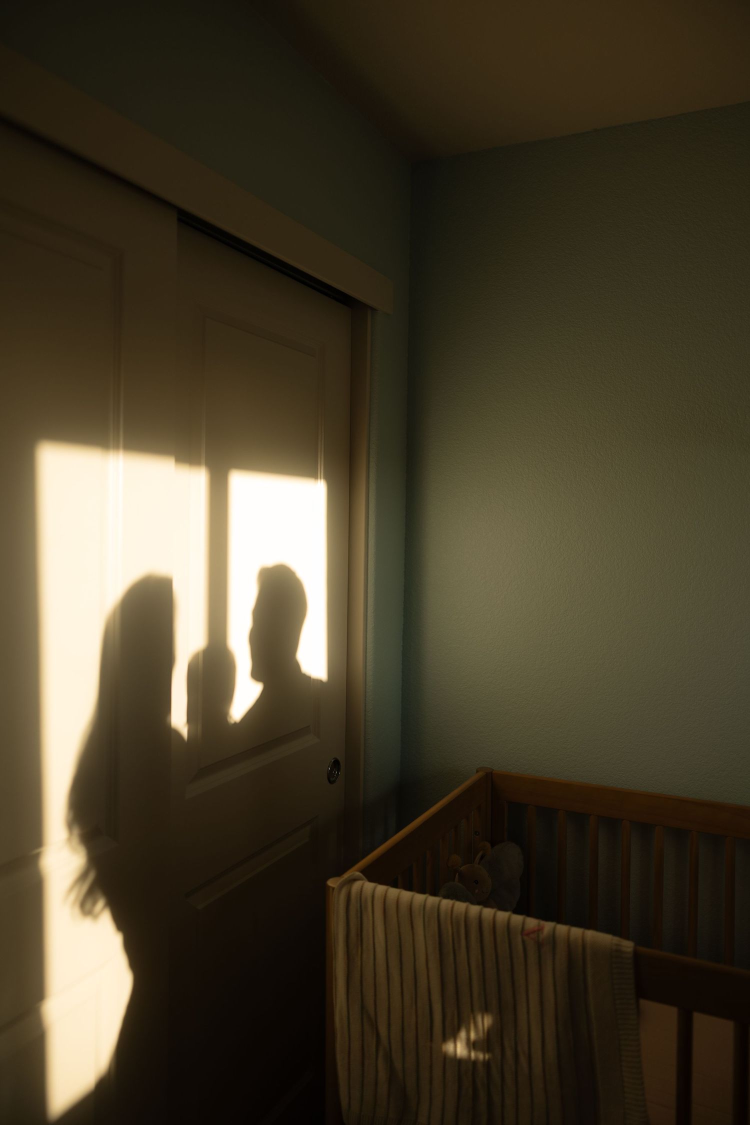 Artistic silhouette of two parents cast on a nursery wall beside a wooden baby crib in warm golden light.
