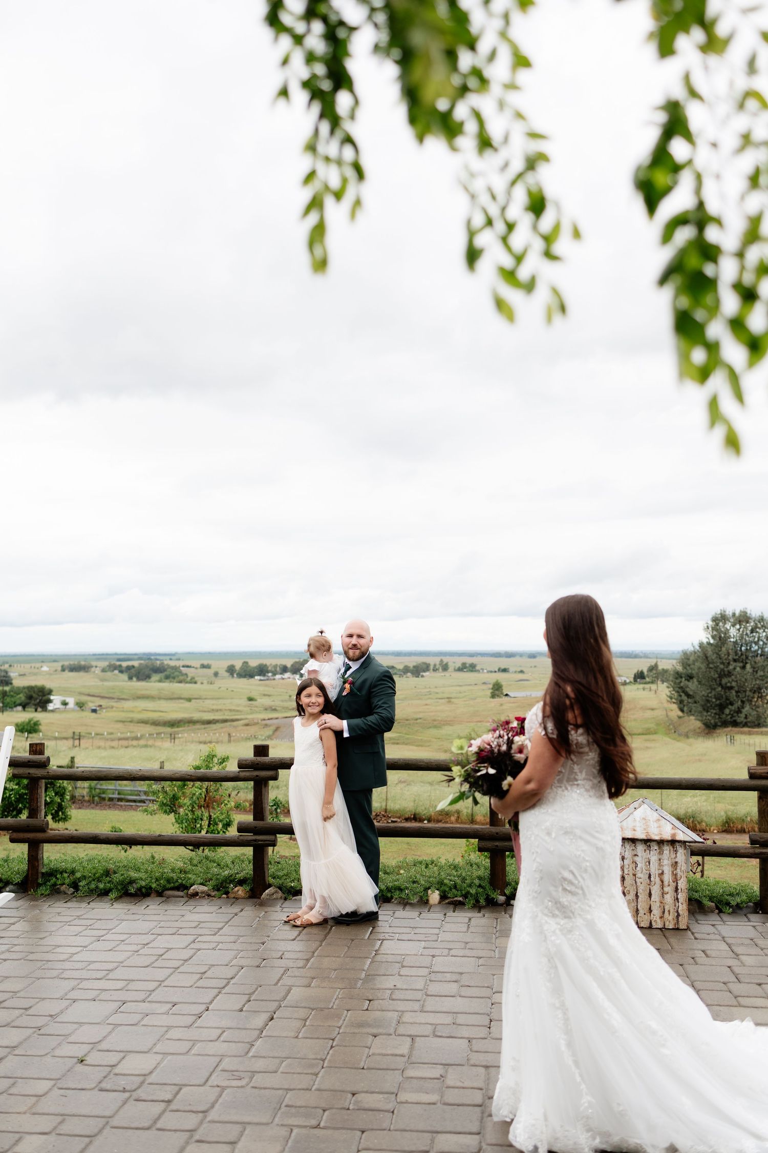 Bride in lace gown holds bouquet while groom lifts child on scenic outdoor terrace.