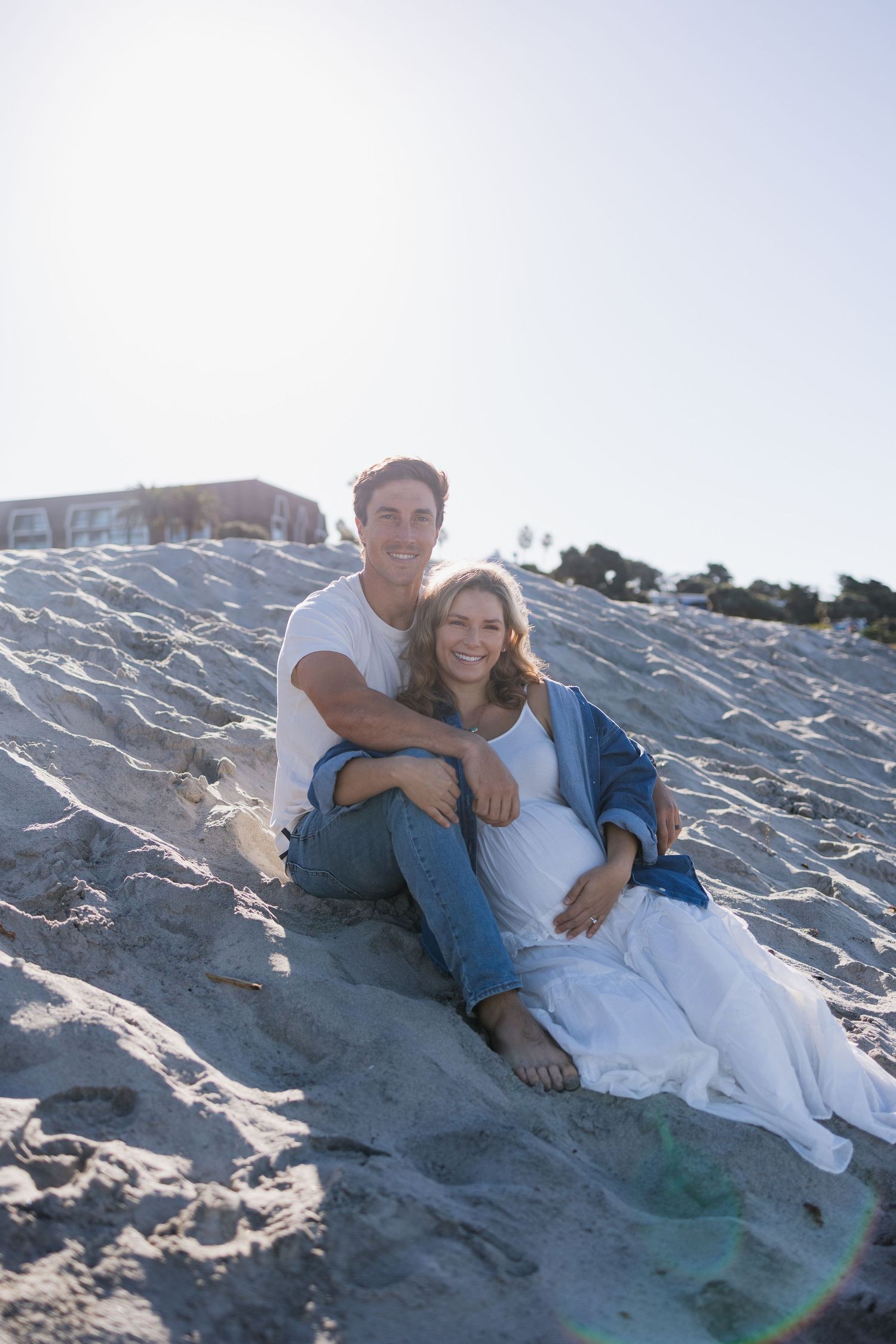 Happy couple sitting together on sandy beach dunes, woman in white dress and blue jacket with visible baby bump.
