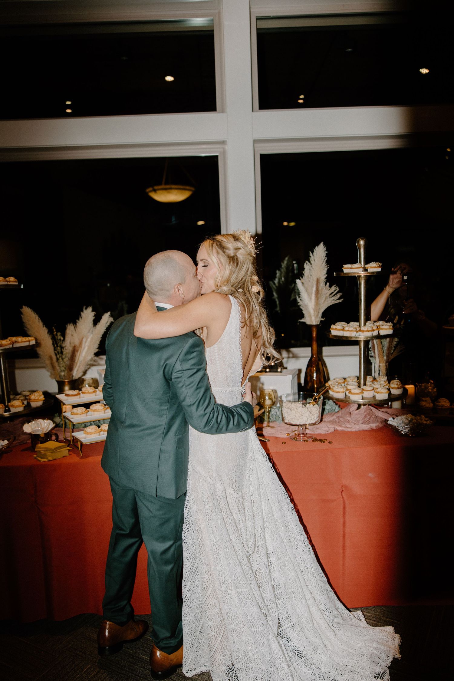 Bride and groom share kiss at dessert table with tiered cake and cupcake display at reception.