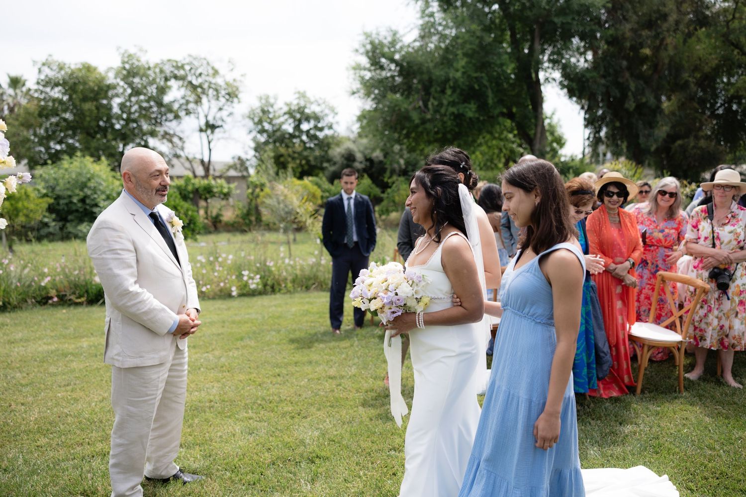 Bride walks down the aisle with a bridesmaid in blue as the groom waits at an outdoor ceremony.