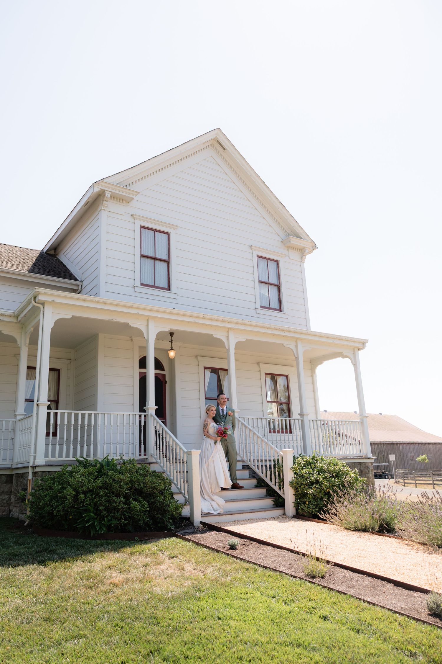 Bride and groom pose on the steps of a charming white farmhouse with a wraparound porch on their wedding day.