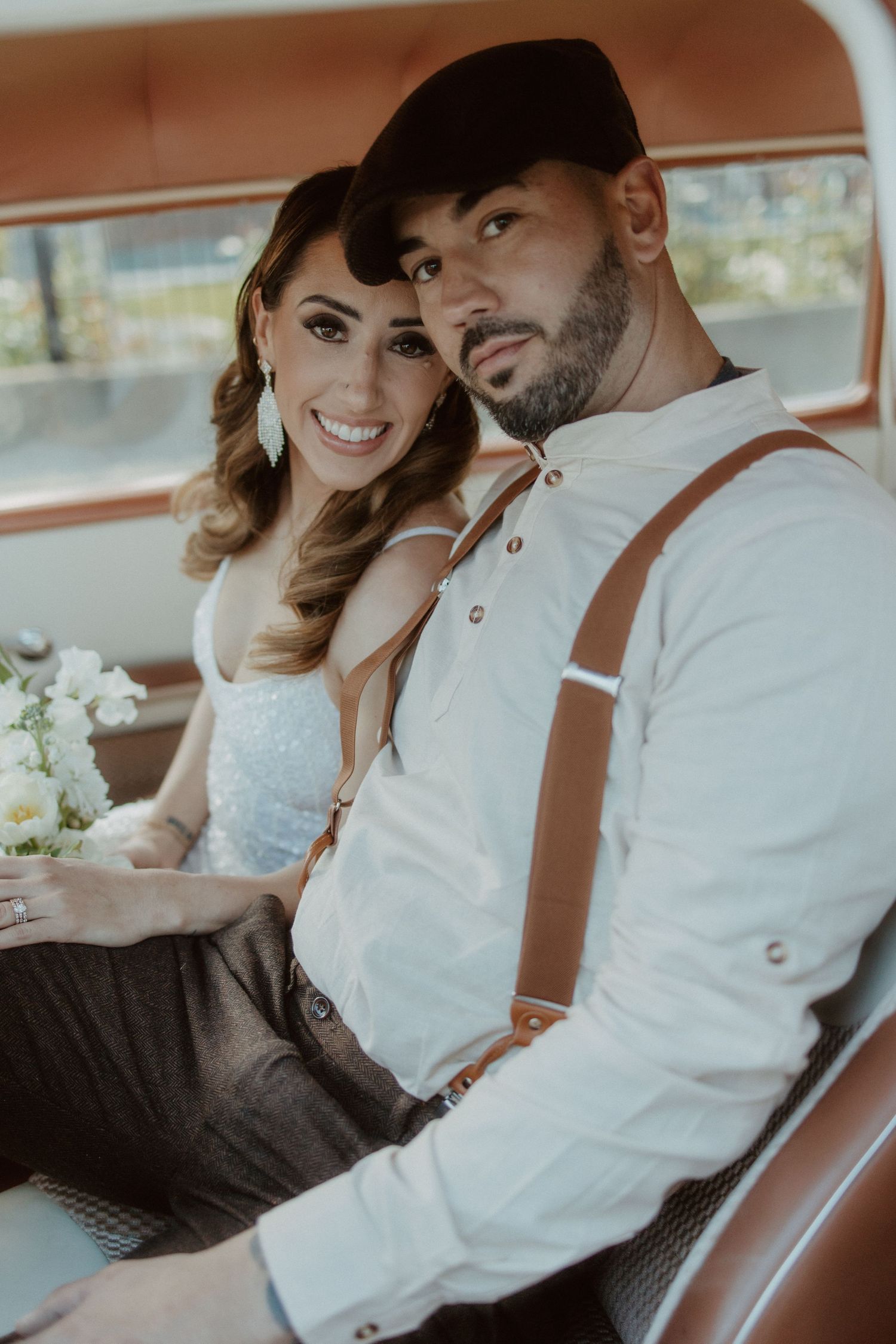 Bride and groom sitting in vintage car, groom wearing suspenders and cap, bride holding white bouquet.