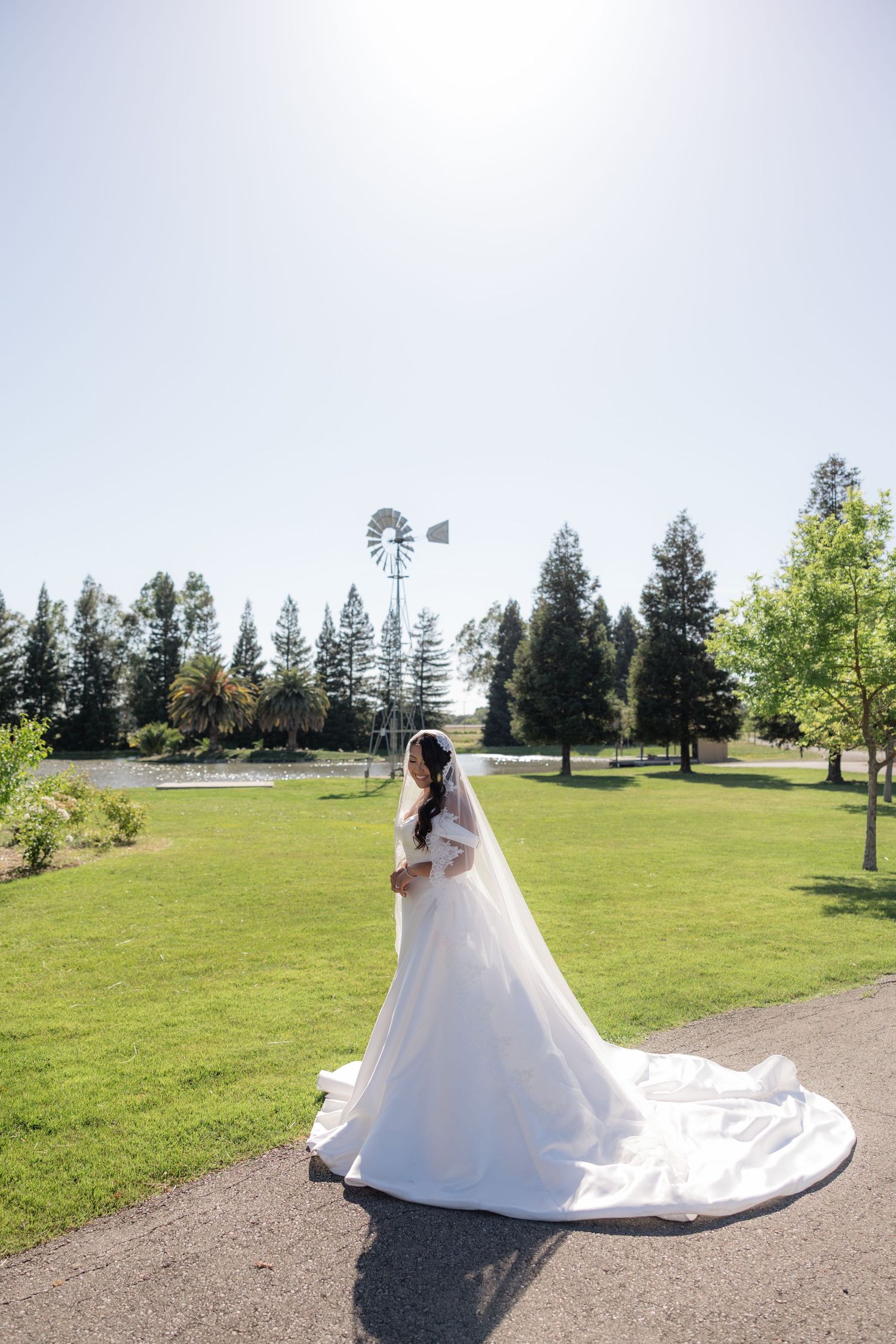 Bride in elegant white gown with long train and veil poses in lush green park with windmill backdrop.