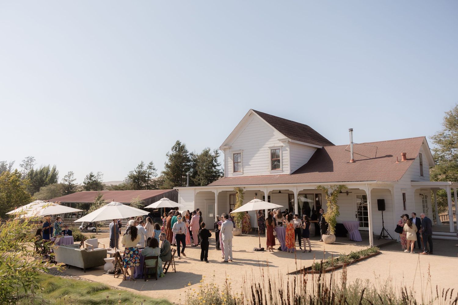 Guests mingle outside a charming white farmhouse venue with patio umbrellas on a sunny day.