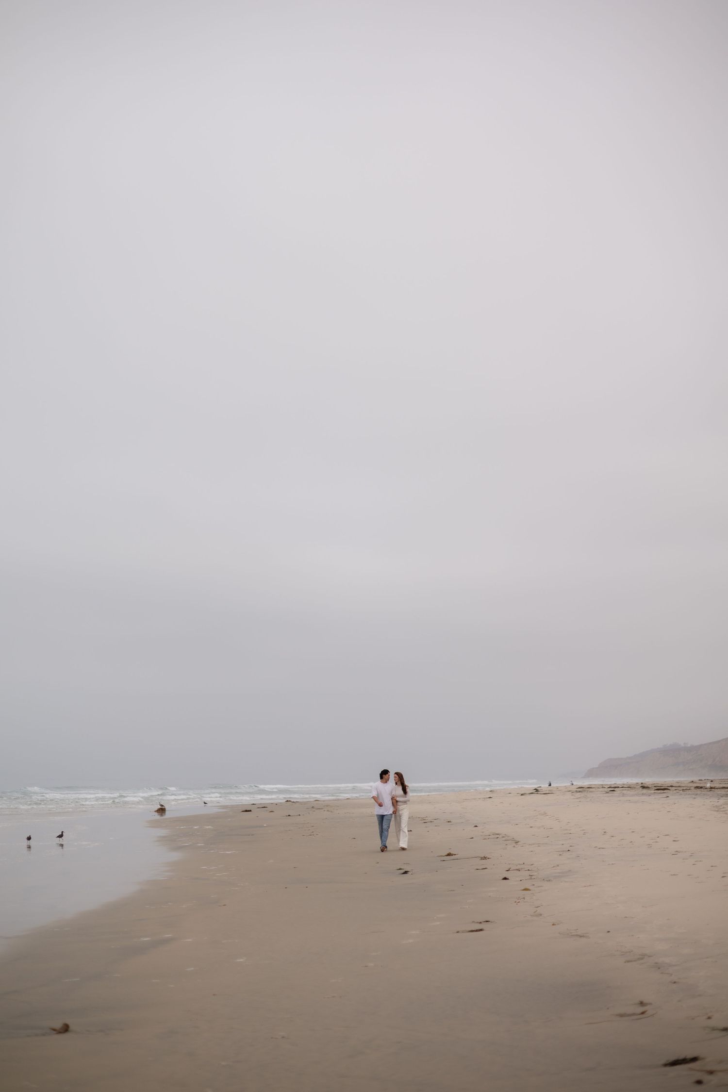 Couple walking on a misty beach with overcast skies, waves, and seabirds on the shoreline.