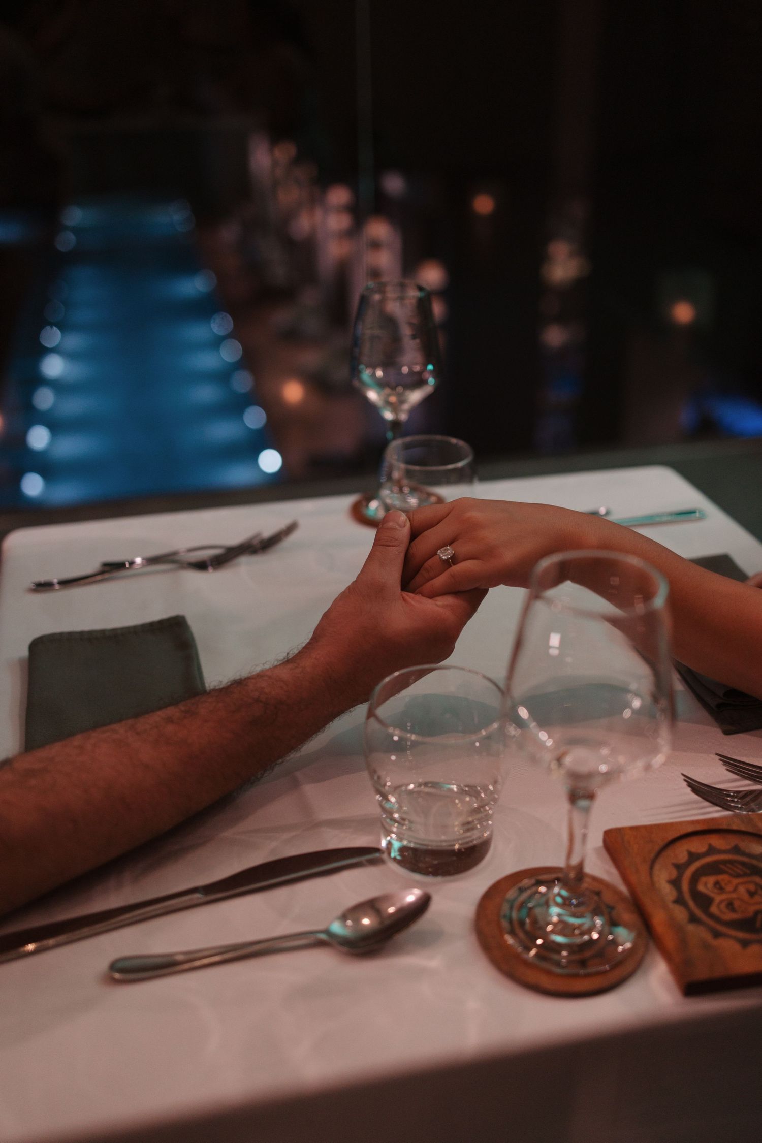 Couple holding hands across a candlelit dinner table with elegant glassware and poolside ambiance.