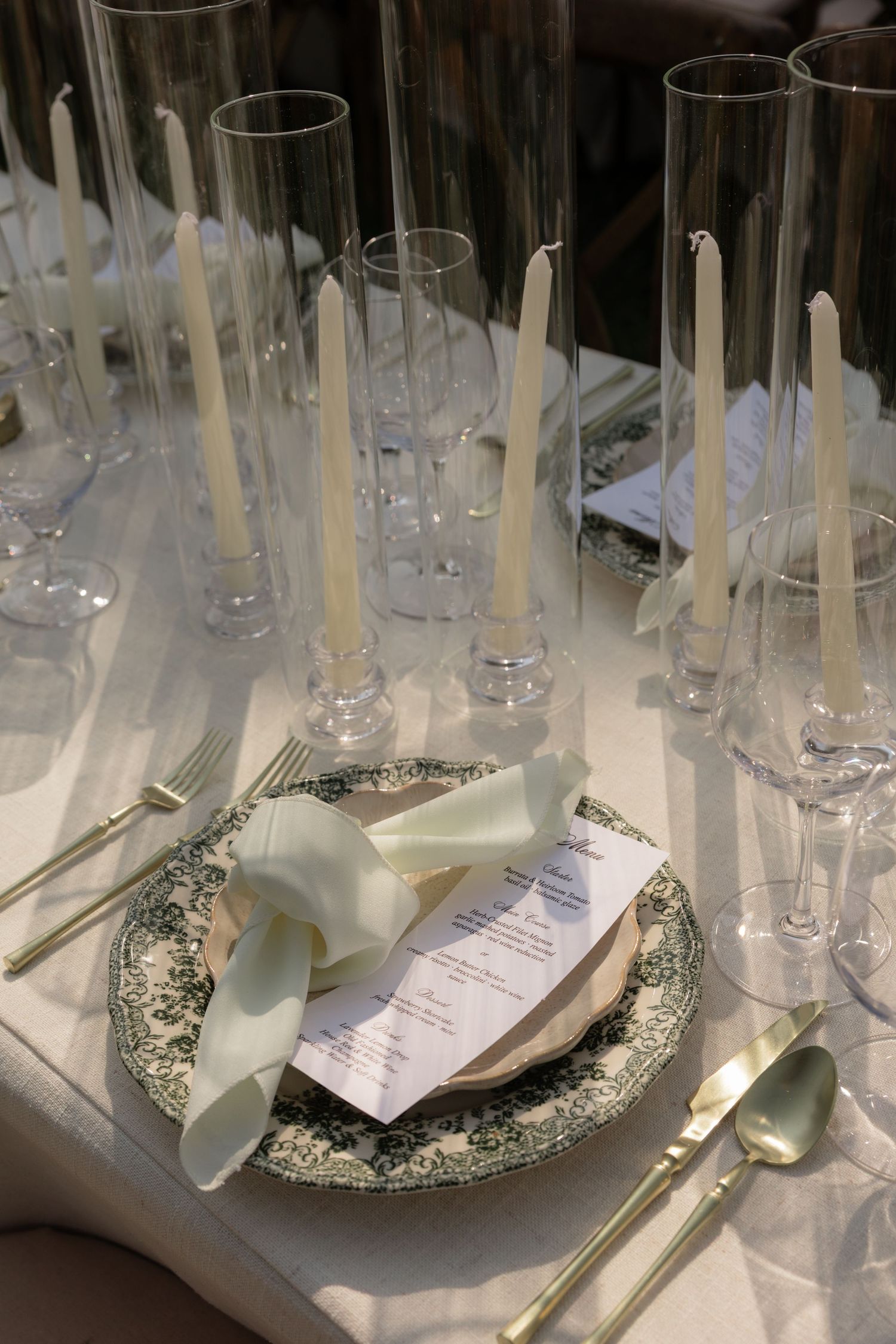 Elegant wedding table setting with white candles, crystal glasses, ornate plates, and a menu card tied with a ribbon.