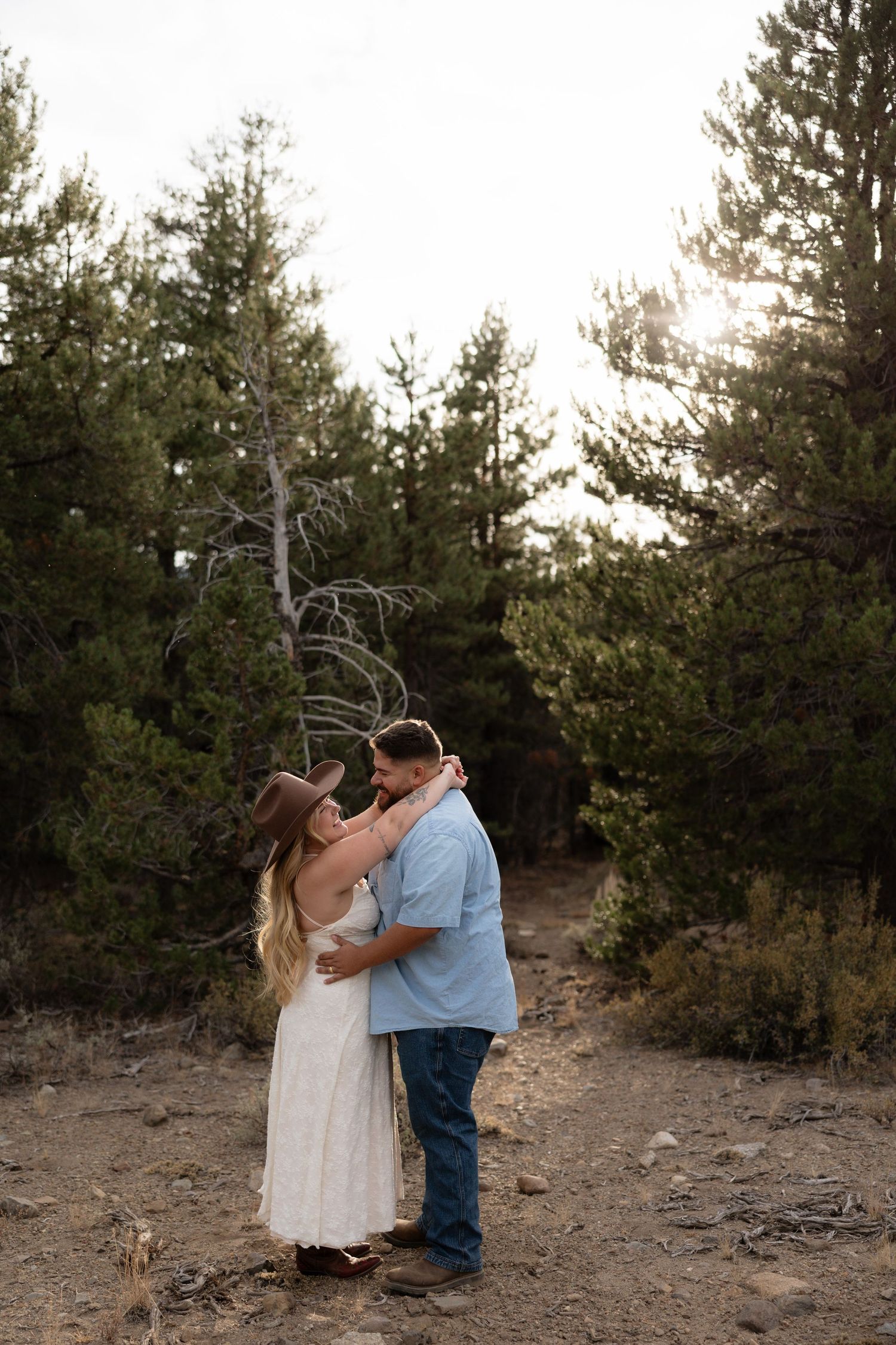 Couple embracing on a forest trail at golden hour, woman in white dress and cowboy hat, man in blue shirt and jeans.