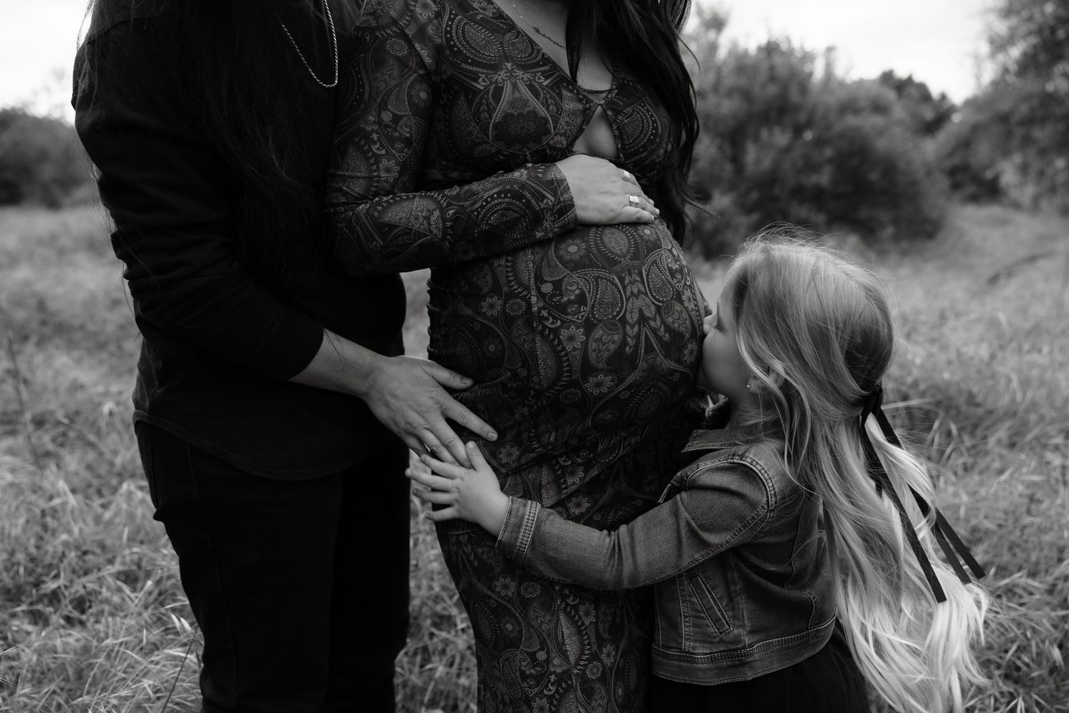 Black and white maternity photo of pregnant woman in lace dress with partner and young daughter outdoors.