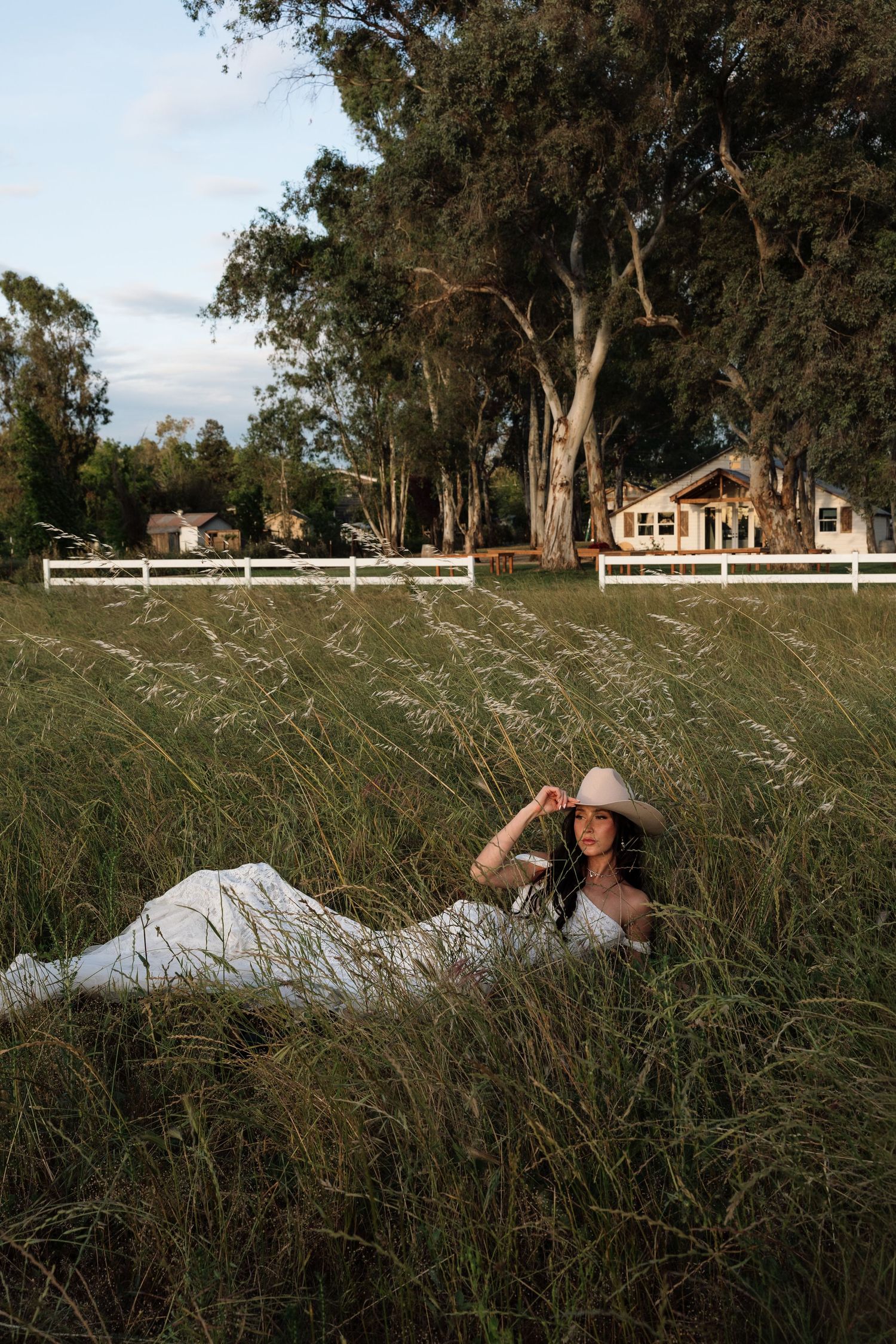 Bride in white dress and cowboy hat lying in tall grass on a rustic farm with white fence and trees.