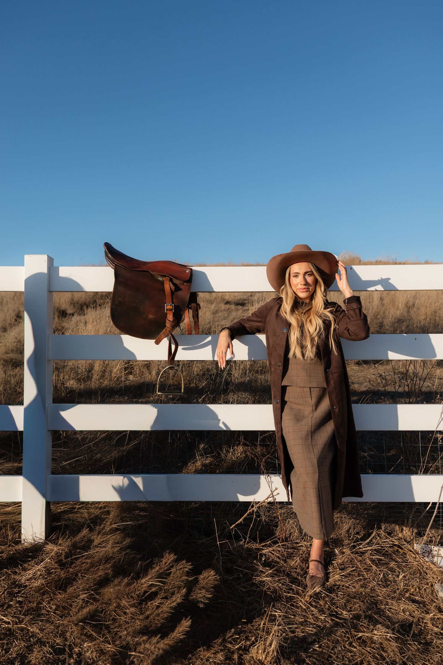 Woman in brown hat and coat leaning against white fence with saddle, rural ranch setting under clear blue sky.