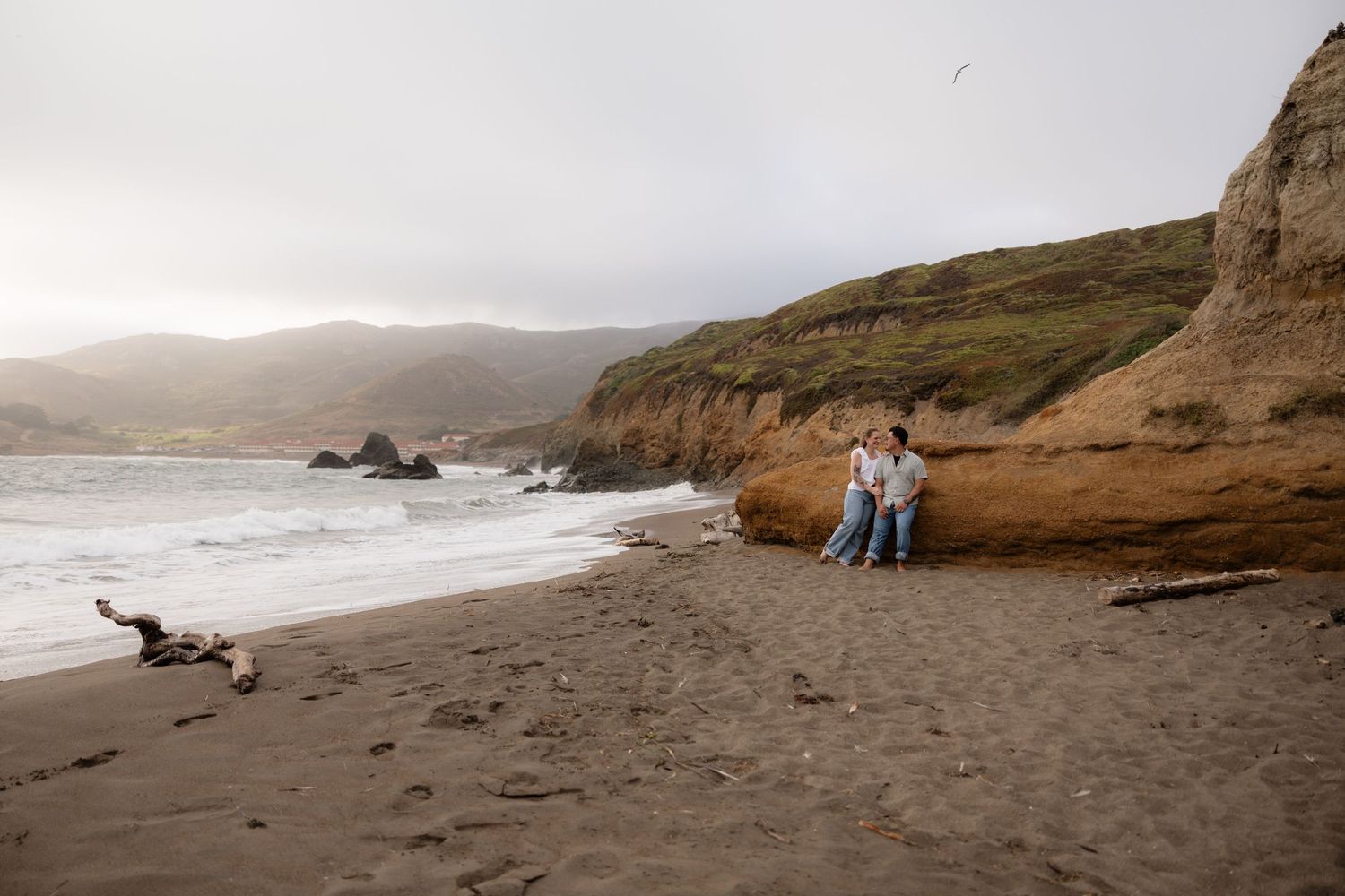 Couple sitting together on a sandy beach with dramatic cliffs and ocean waves in the background.