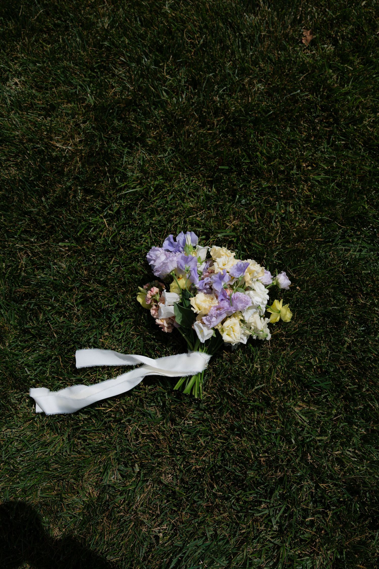 Bridal bouquet of purple, lavender, and white flowers with white ribbon resting on lush green grass.