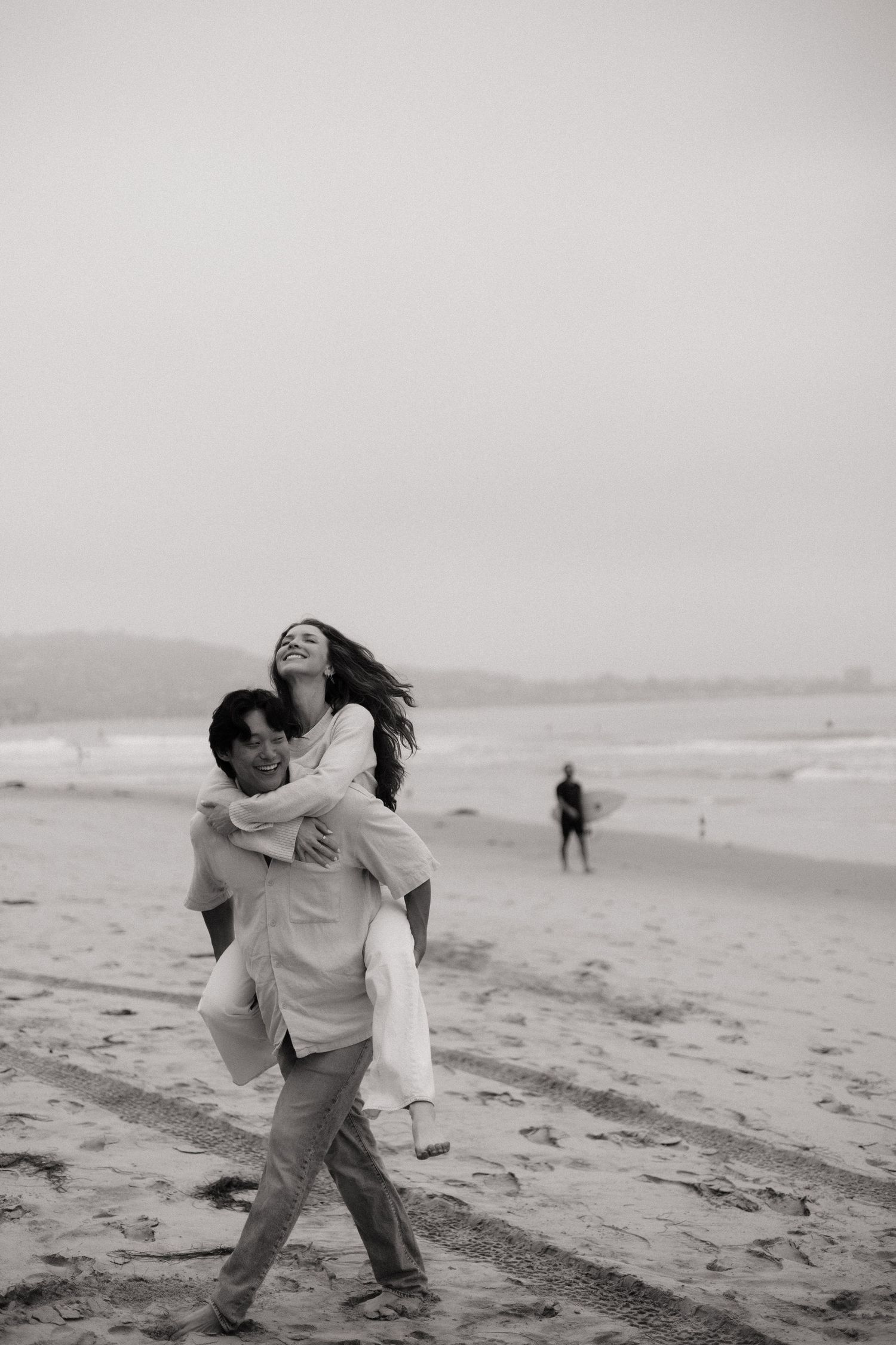 Black and white photo of a man giving a woman a piggyback ride on a beach, both laughing joyfully.