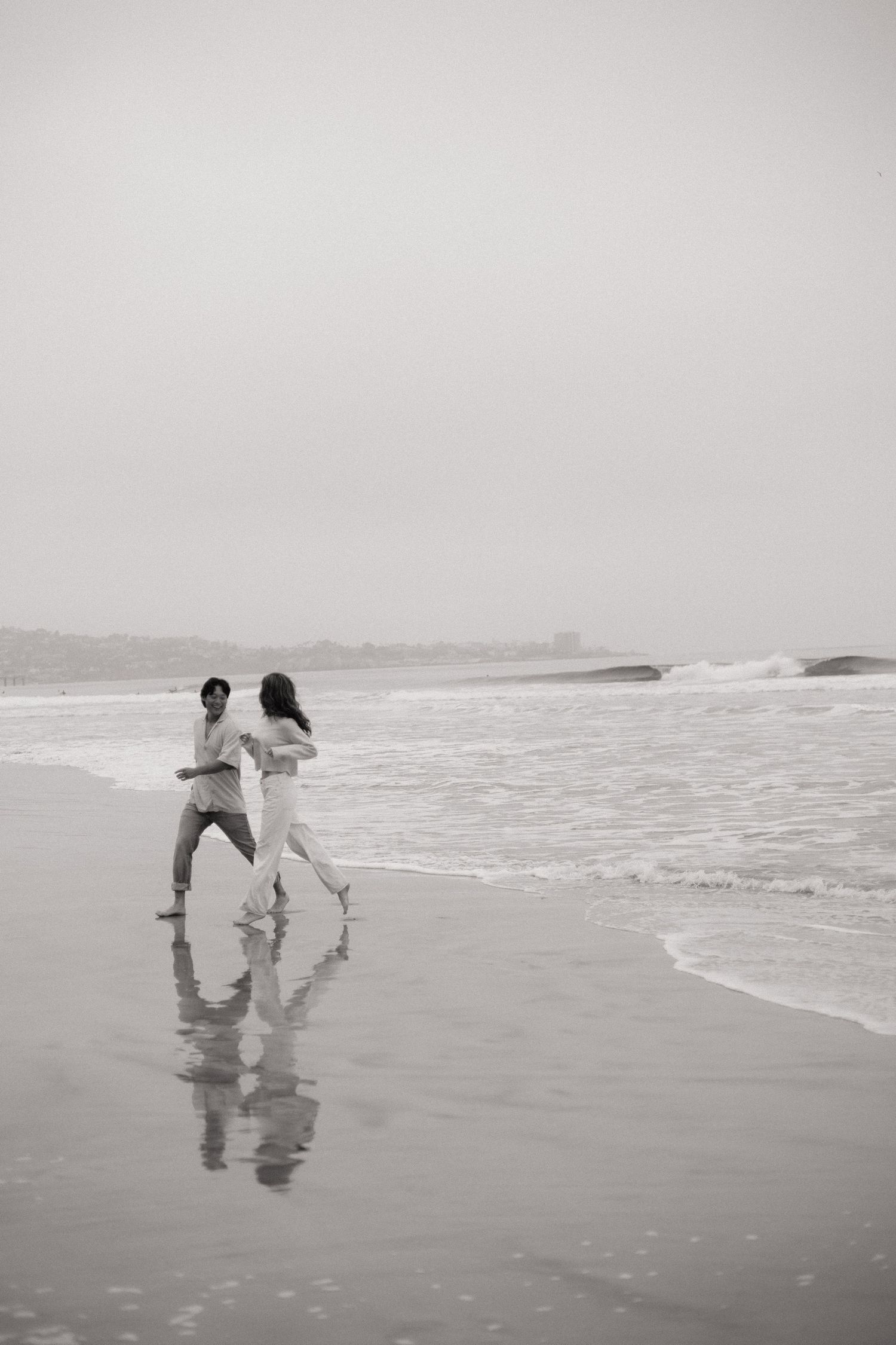 Black and white photo of a couple running along the ocean shoreline, waves crashing beside them.