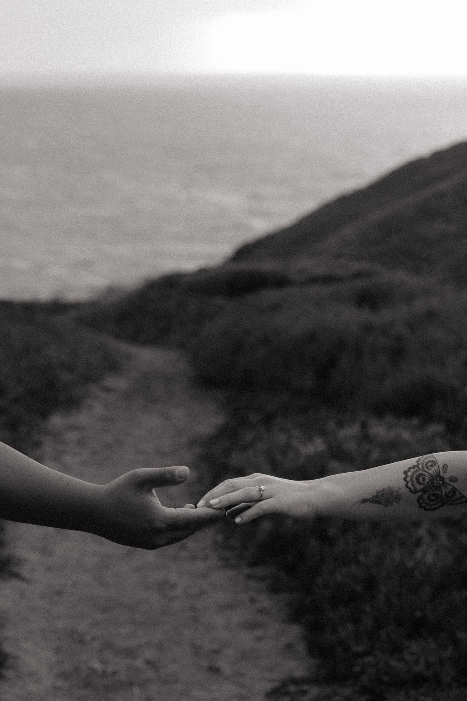 Black and white photo of two hands almost touching on a coastal path, one with a floral tattoo and ring.