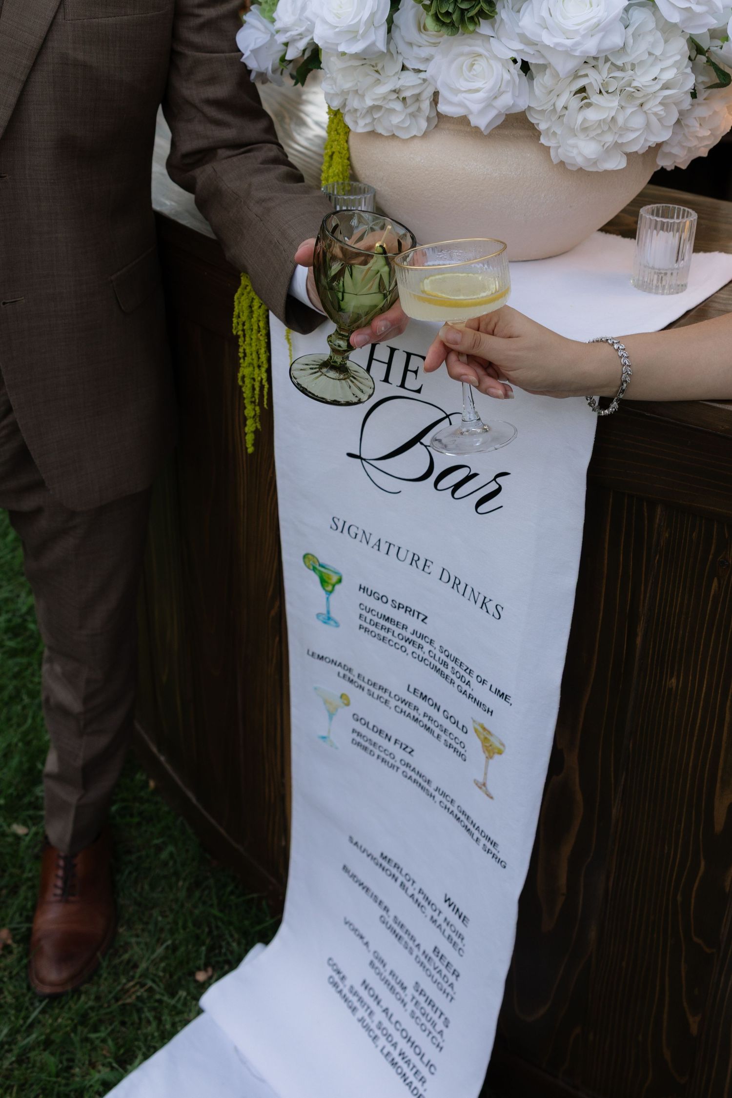 Elegant wedding bar menu sign with signature cocktails, held by guests in formal attire at an outdoor reception.