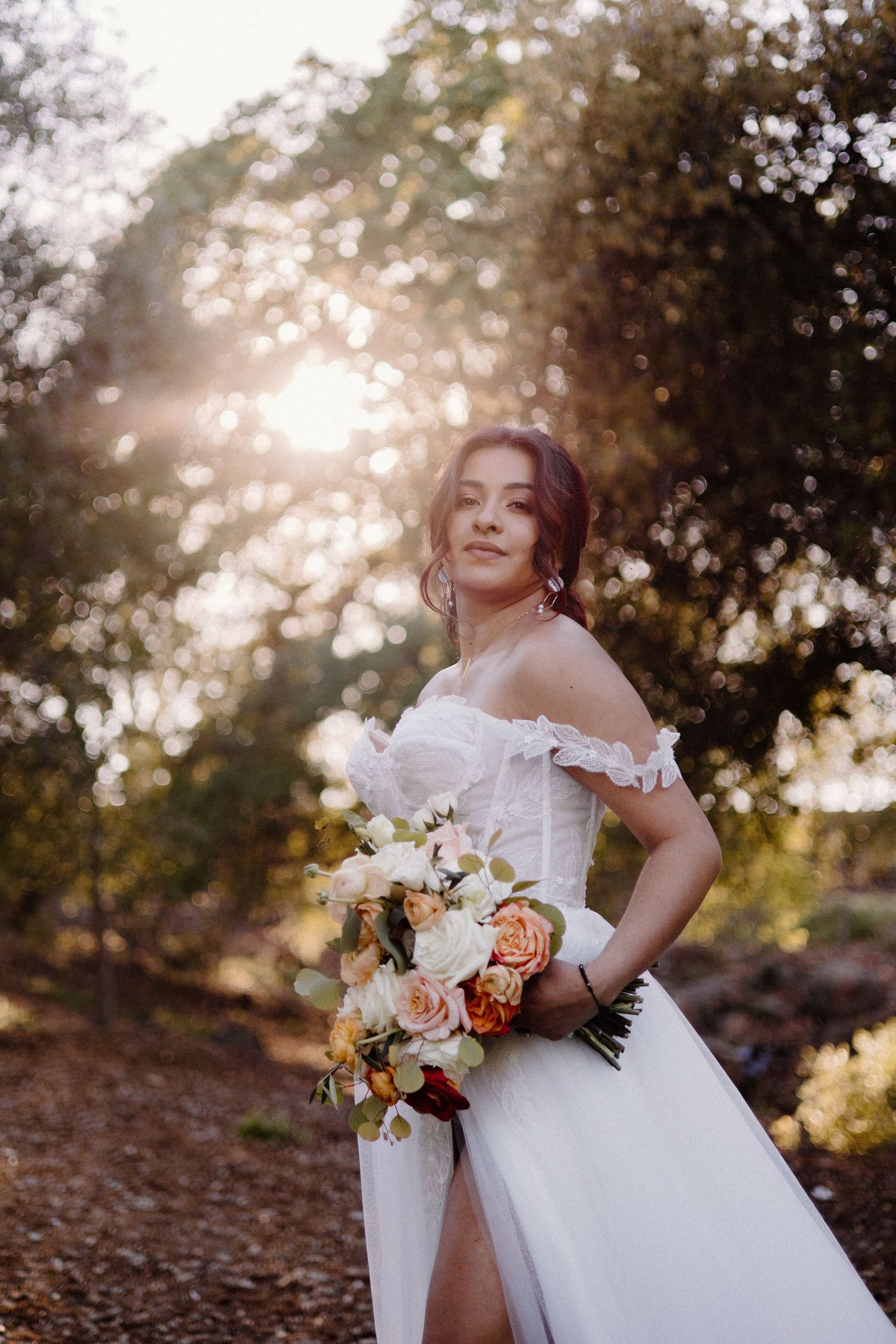 Bride in off-shoulder white gown holding peach and white bouquet in sun-dappled forest setting.