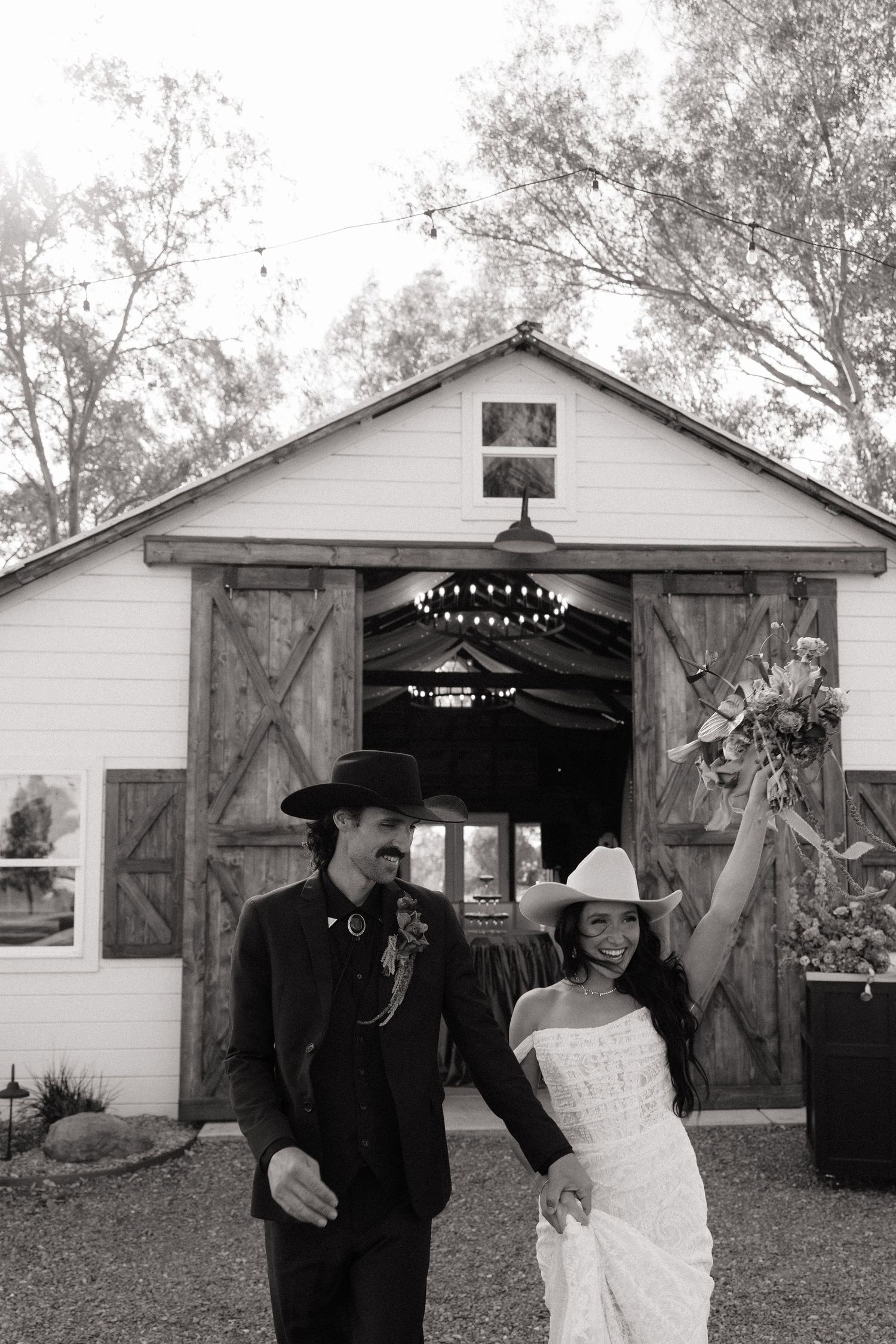 Black and white photo of happy couple in cowboy hats celebrating outside rustic barn wedding venue.