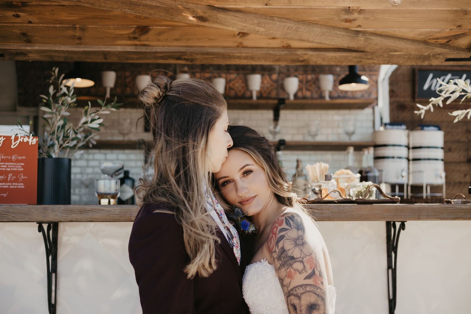Two brides share a tender embrace near a rustic bar setup at their intimate wedding celebration.