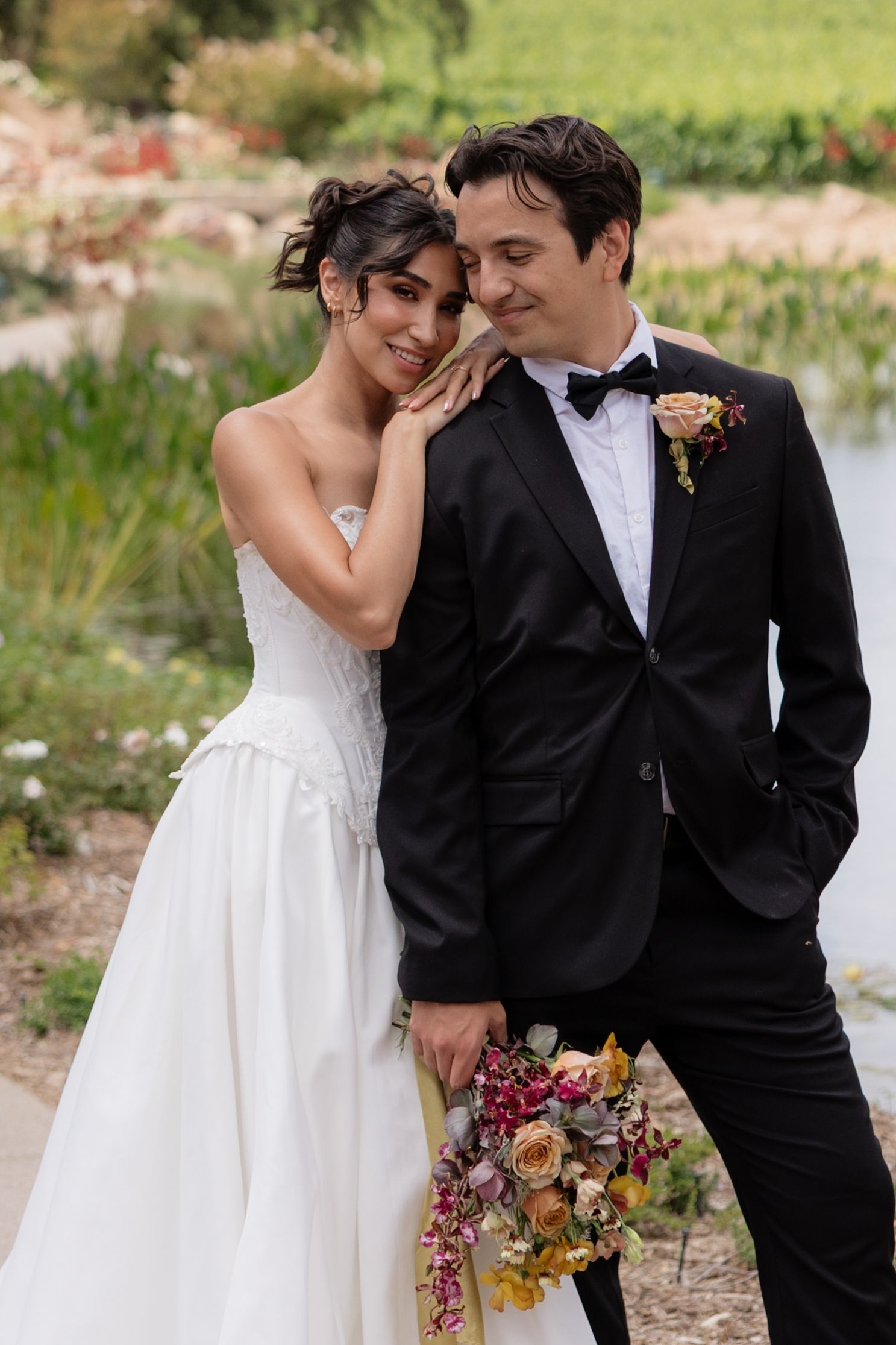 Bride leans on grooms shoulder holding colorful bouquet while posing near a garden pond.