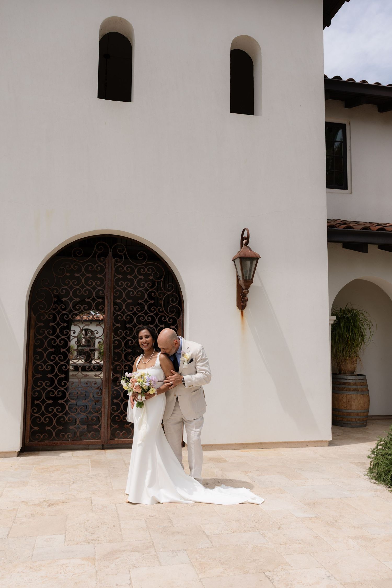 Bride and groom embrace in front of ornate arched iron doors of a white Spanish-style wedding venue with barrel accents.