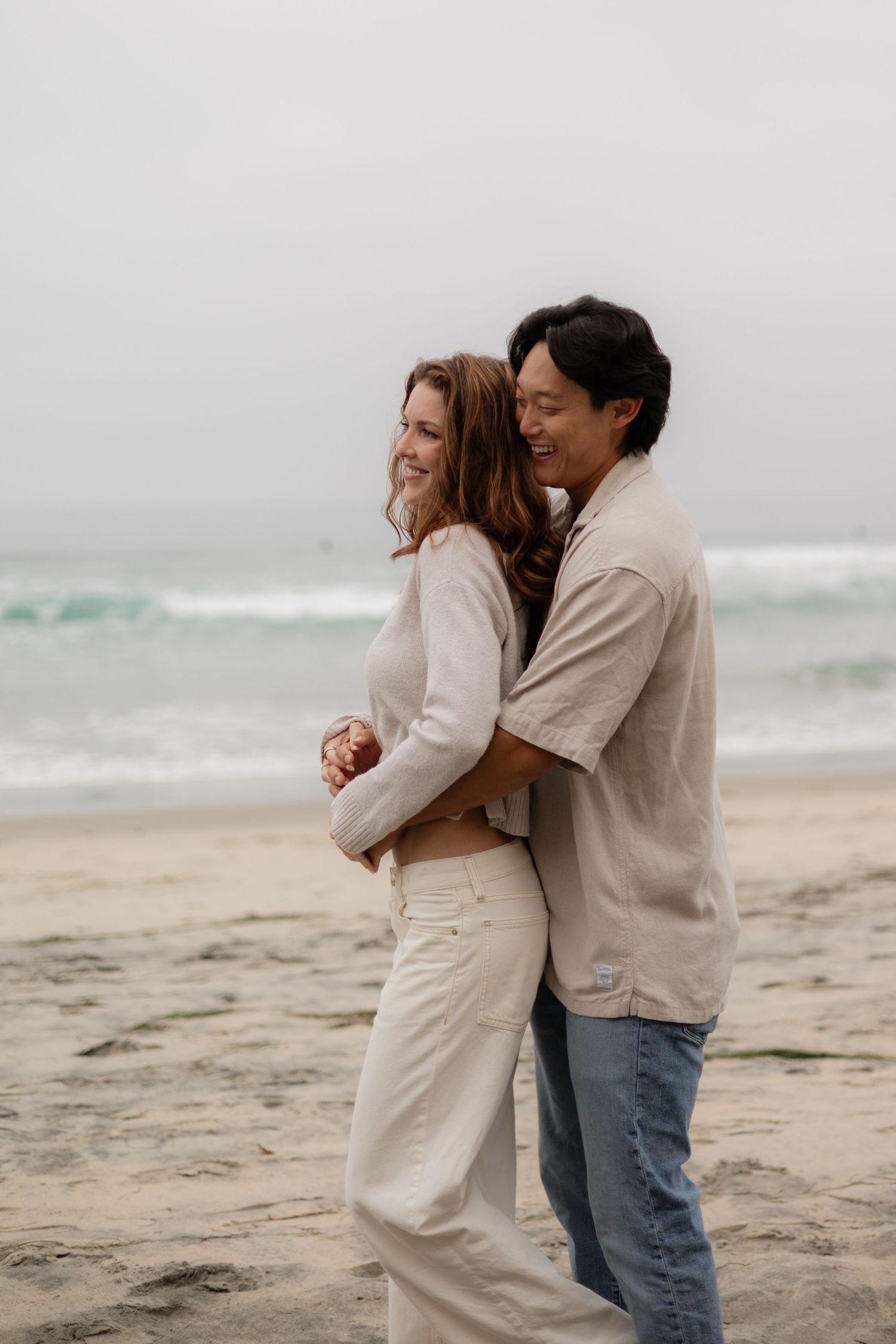 Happy couple embracing on a sandy beach, smiling together with ocean waves in the background on a cloudy day.