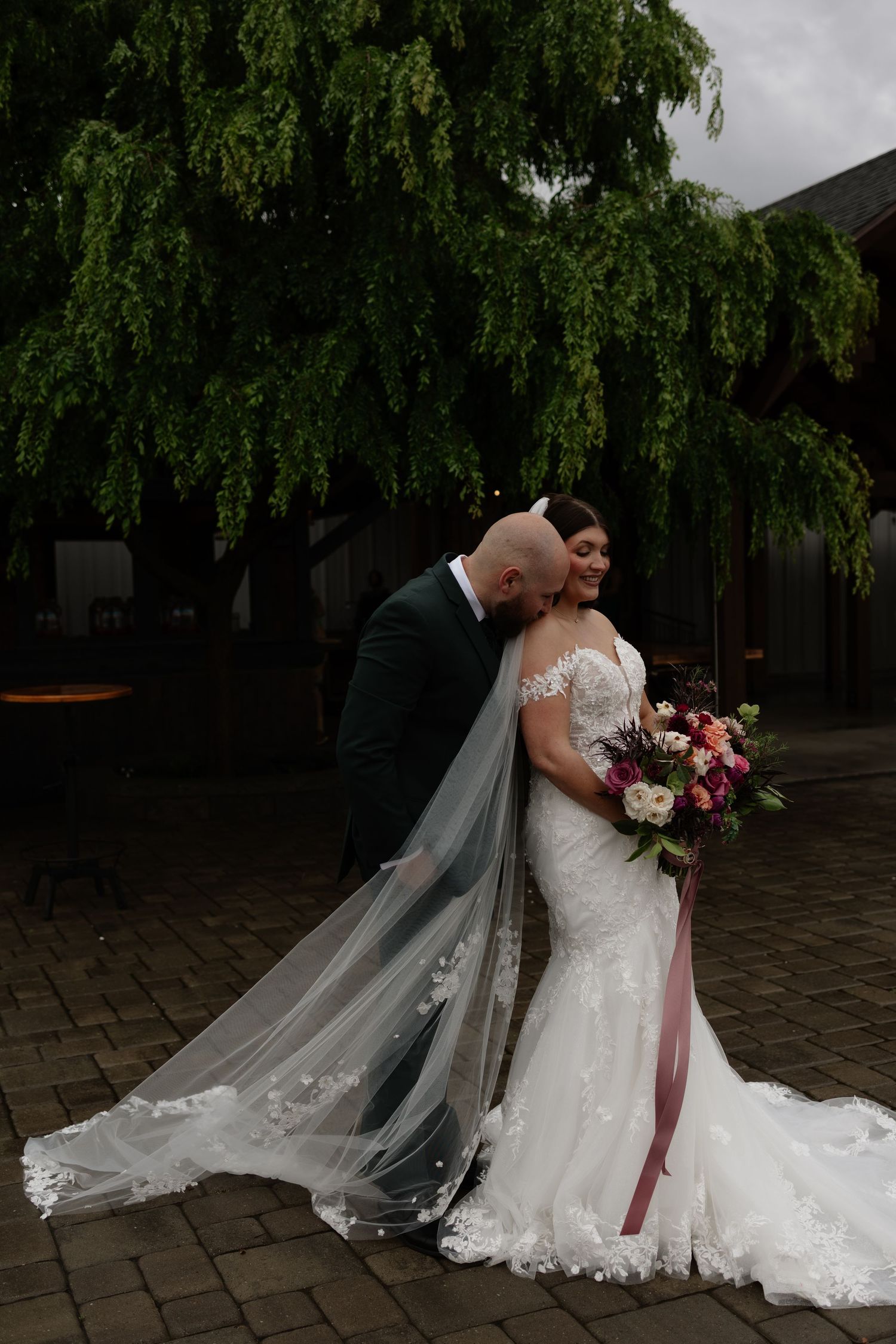 Groom kisses bride in lace mermaid gown beside weeping willow, her floral bouquet trailing.
