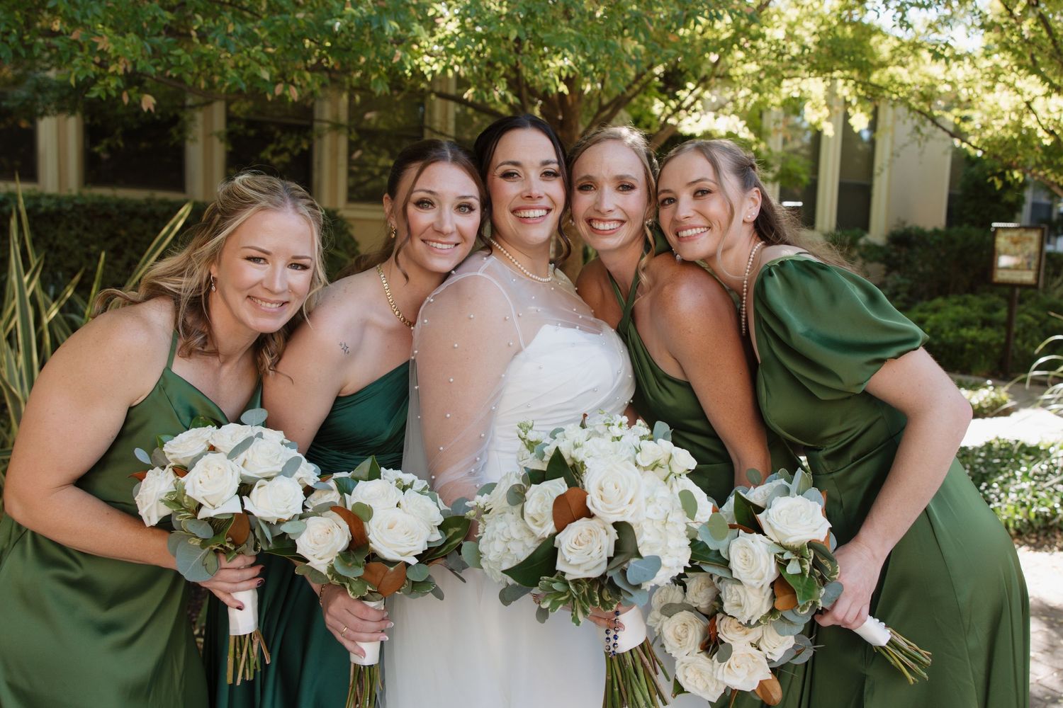Bride in white gown with three bridesmaids in green dresses holding white and green bouquets outdoors.